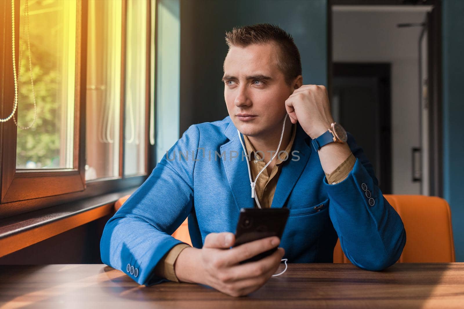 A handsome businessman of European appearance stylish portrait, holds a phone in his hand and listens to music in headphones sitting at a table in a cafe.