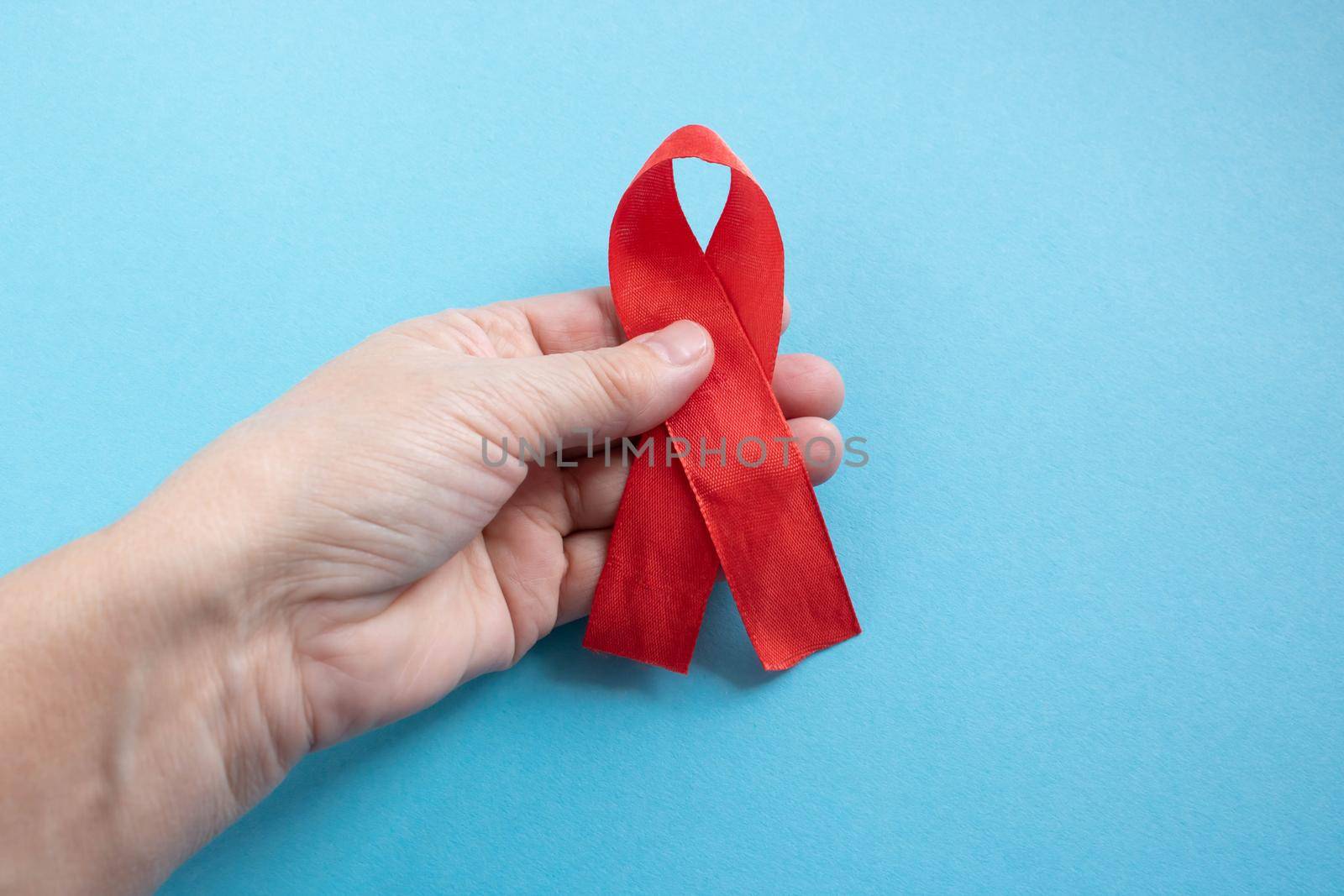 A hand holds a red ribbon on a blue background - the concept of World AIDS Day, Donor Day, Hemophilia Day.