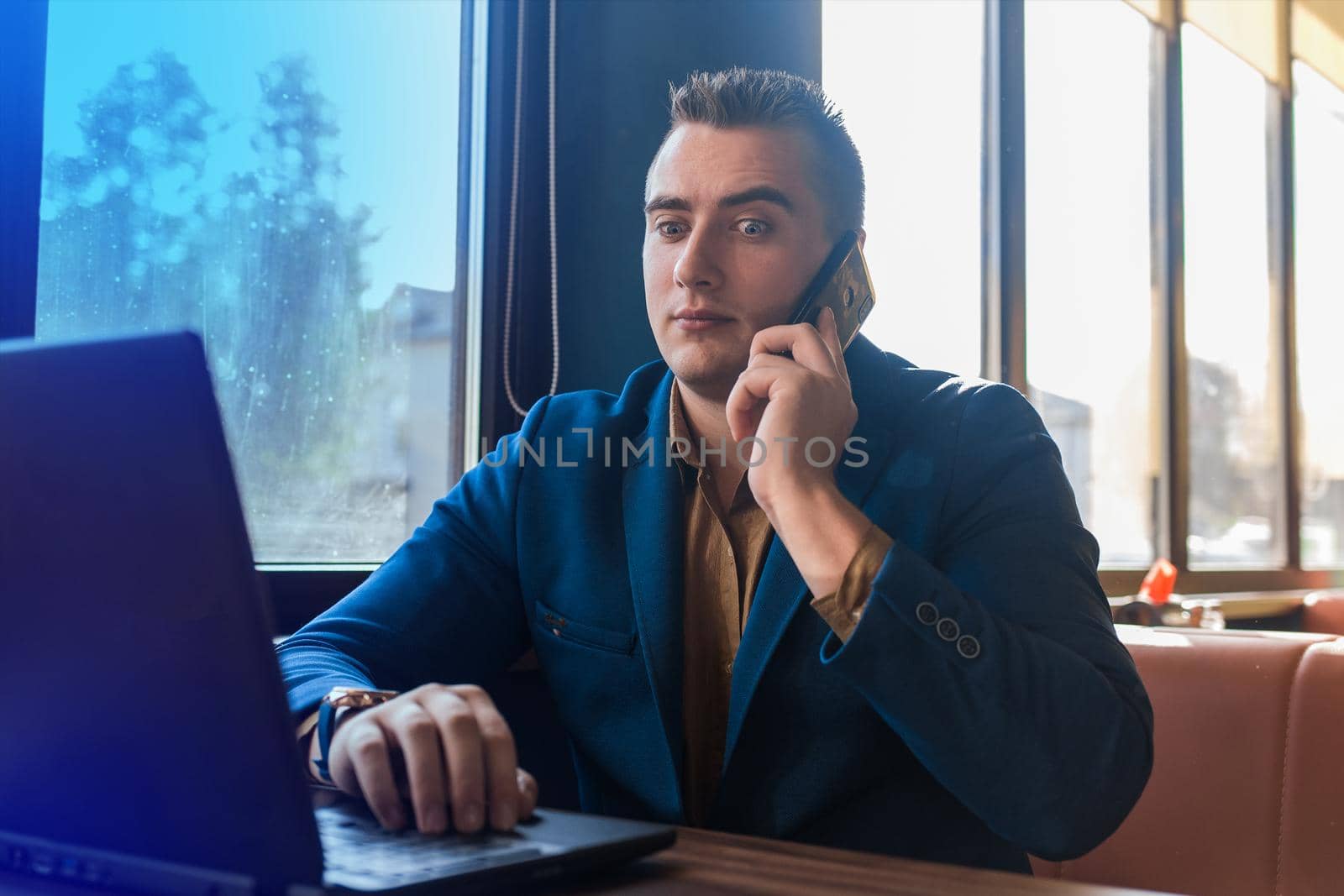 A business man surprised businessman in an attractive European-looking suit works in a laptop, talks on a cell or mobile phone, sitting at a table in a cafe by the window.