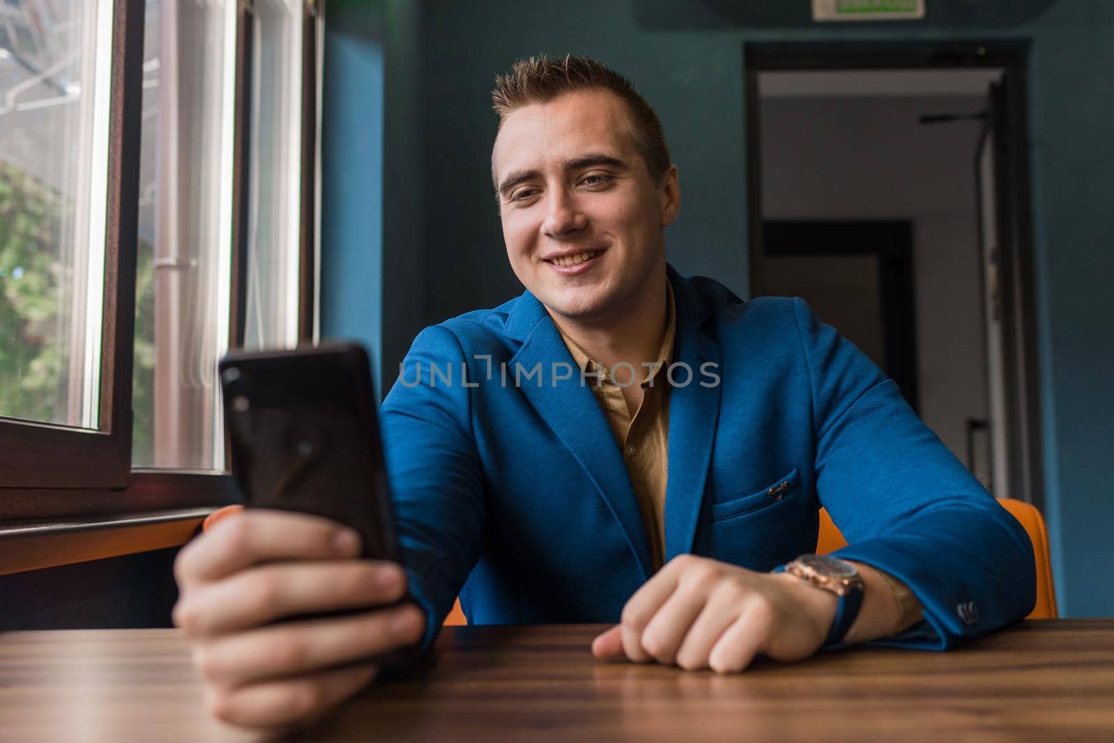 A positive, smiling young guy of European appearance, a stylish businessman, looks at a mobile phone or smartphone and takes a selfie or talks via video link.