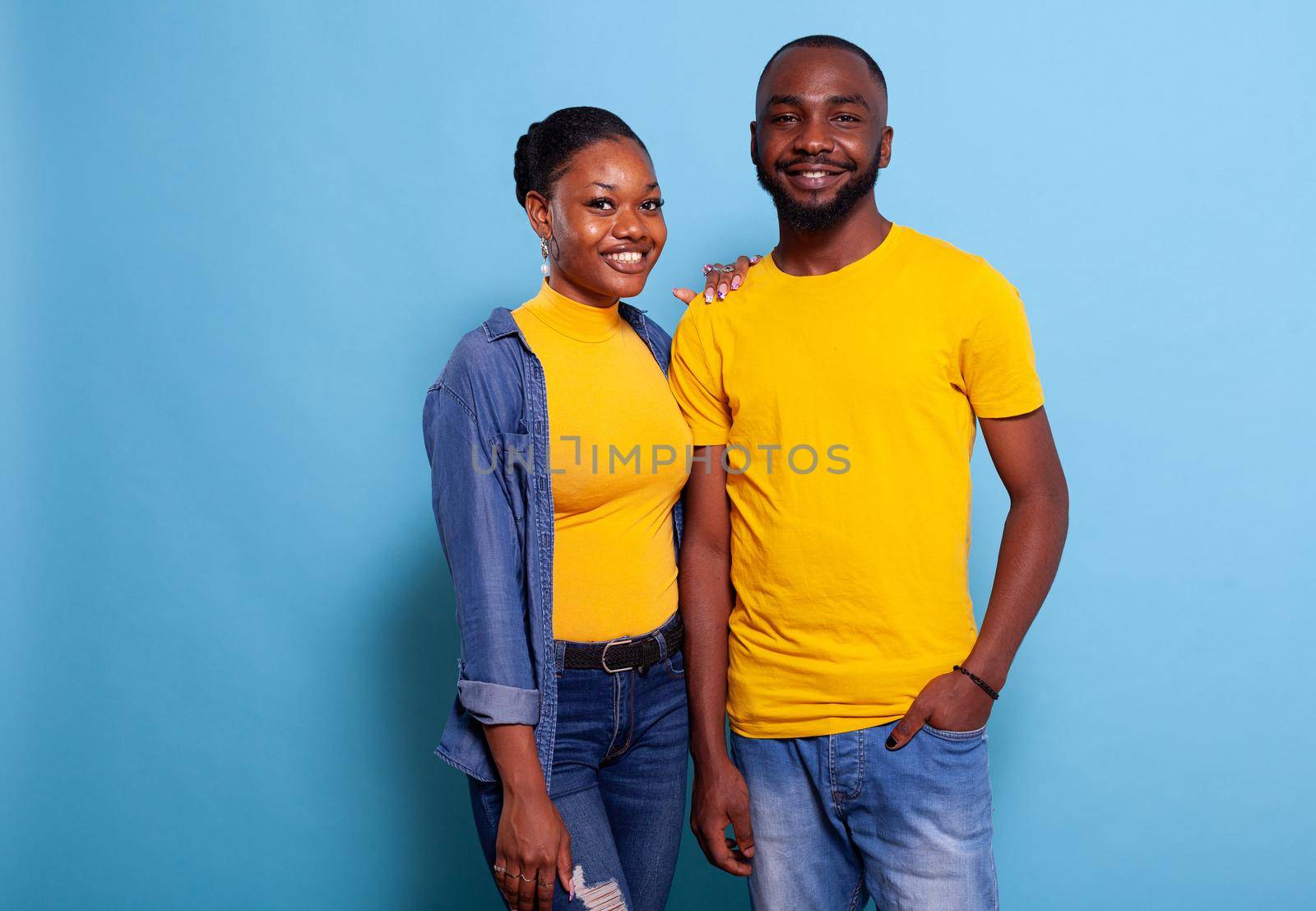 Smiling couple standing in embrace over blue background. Man and woman in love expressing love and affection with hug in studio. Lovers feeling happy and peaceful, looking at camera.