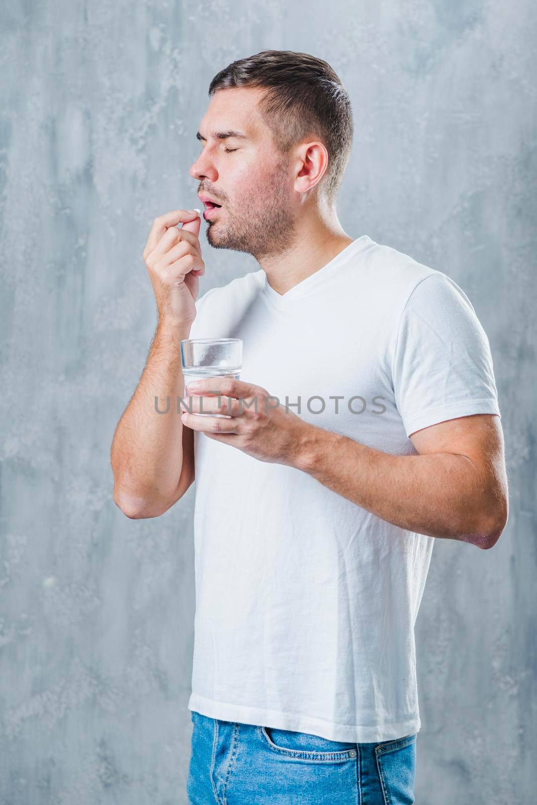 sick young man standing against gray backdrop holding water glass hand taking pill by Zahard