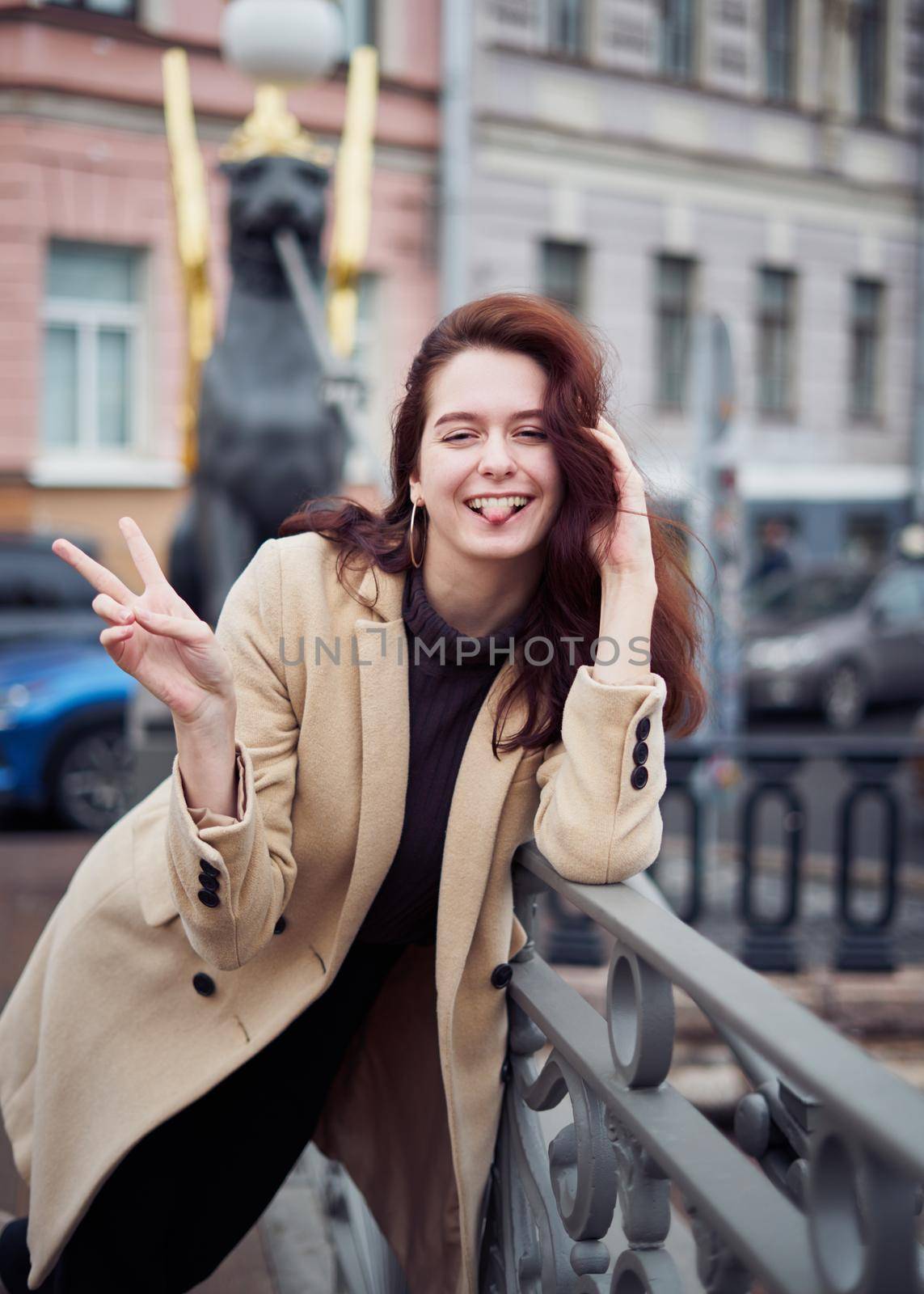 Beautiful serious stylish fashionable smart girl standing on bridge, smiling, show tongue, show victory sign with hand. St. Petersburg city. Charming funny happy woman with long dark hair, vertical