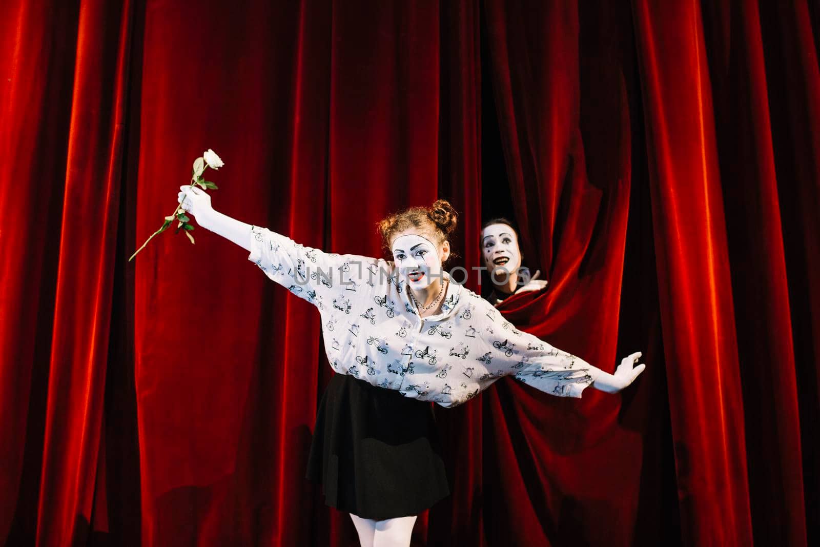 female mime holding white rose performing stage with male mime peeking curtain