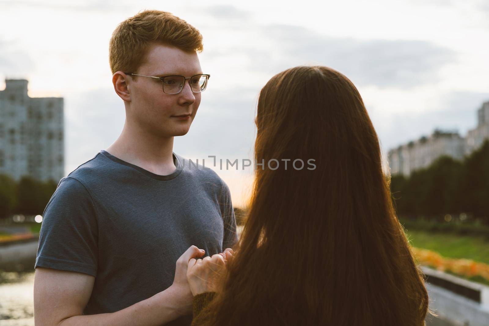 Girl with long thick dark hair holding hands redhead boy in blue t-shirt on bridge, teen love at evening. Boy looks tenderly at girl, young couple. Concept of teenage love and first kiss, sincere feelings of man and woman, city, waterfront. Close up by NataBene