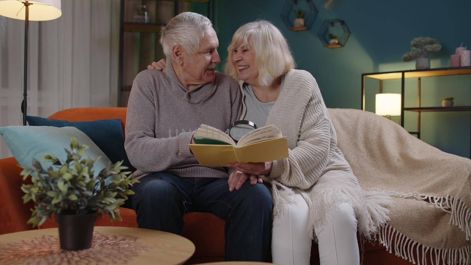 Happy senior grandfather, grandmother relaxing talking enjoying leisure hobbies together at evening home, elderly husband grandpa reading book with old wife granny use magnifying glass sitting on sofa