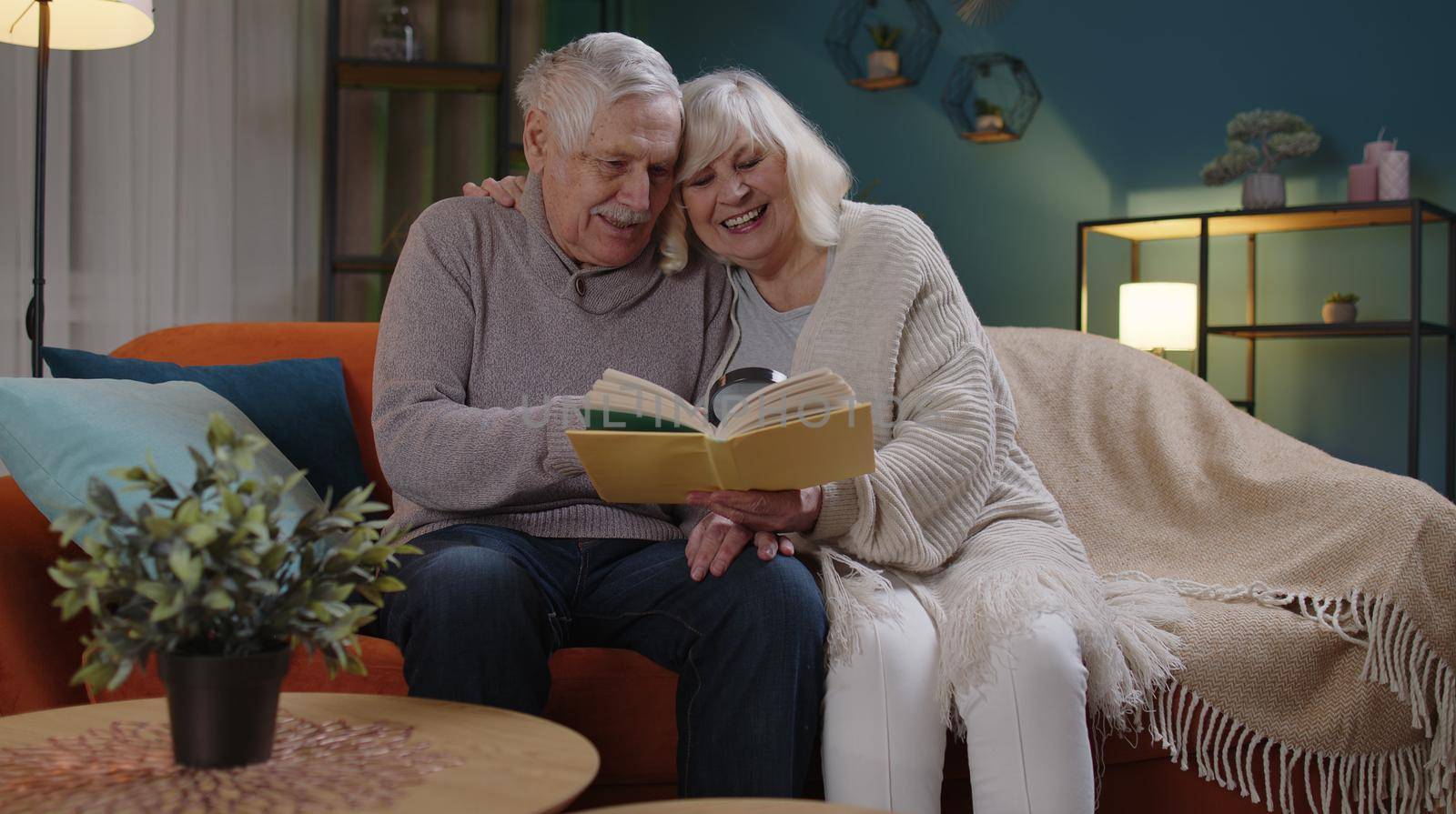 Senior grandparents couple relaxing, reading book, talking enjoying leisure hobbies at night home by efuror