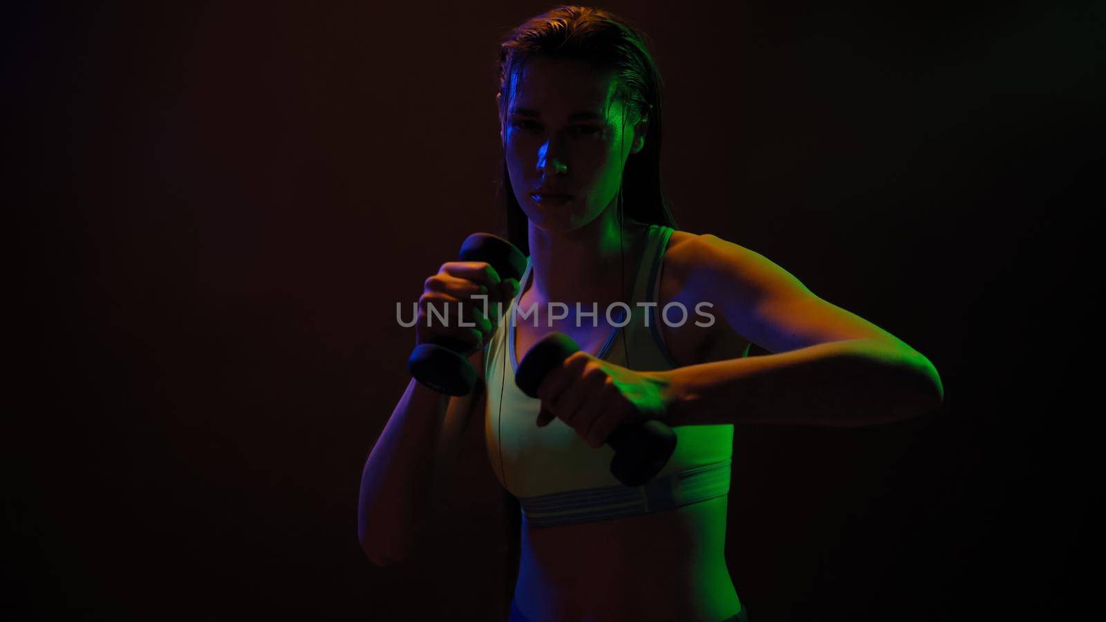 Sportive girl posing doing hand workout biceps with dumbbells standing in neon lights wearing sport top working on a arm weight hit isolated on black background. Neon light concept.
