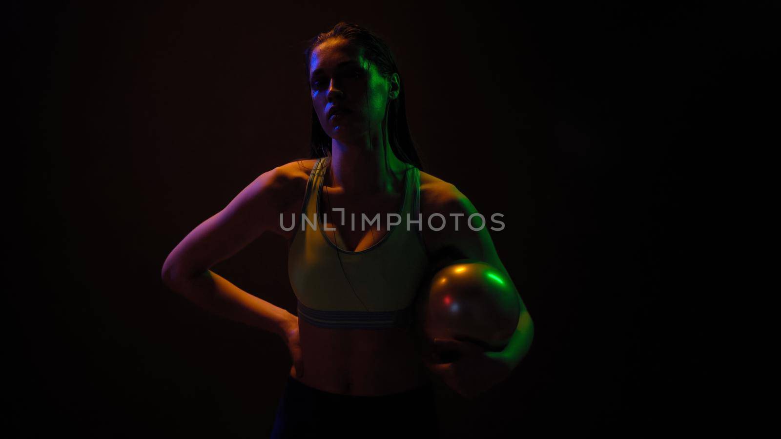 Professional athlete girl posing with power exercise ball standing in neon lights wearing sport top working on a weight fitness isolated on black background. Neon light concept.