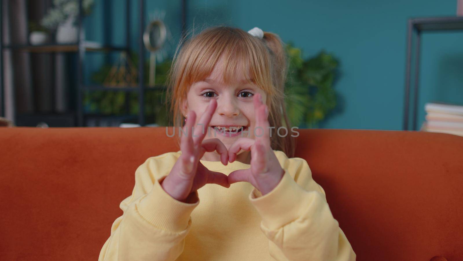 Portrait of funny Caucasian little smiling child girl kid sitting on sofa at home looking at camera makes heart gesture demonstrates love sign expresses good feelings, sympathy. Carefree children