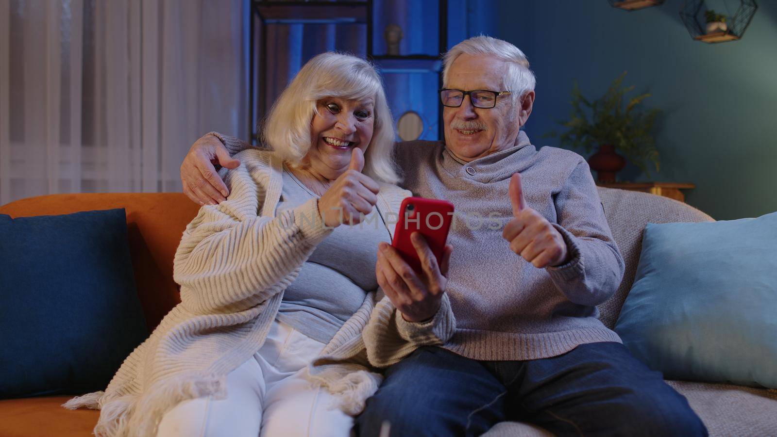 Happy senior elderly couple grandfather and grandmother looking on mobile phone making online shopping and smiling. Mature family husband wife watching news videos, using social media at home