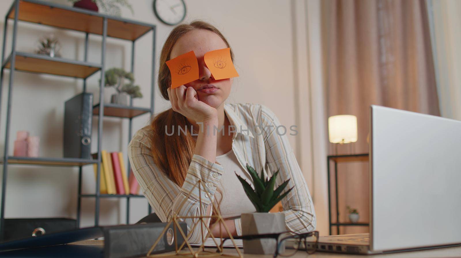 Tired woman freelancer using laptop computer napping at home office desk with funny stickers on eyes sleeps at workplace, suffering migaine headache after e-learning, remote working. Employee burnout