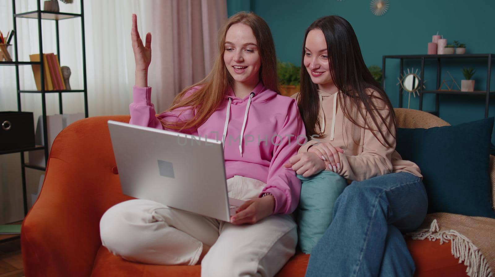 Cheerful girls friends siblings watching photos on laptop, online dating app. Happy two female women couple family on sofa at home, enjoy chatting, spend leisure time on notebook social media network