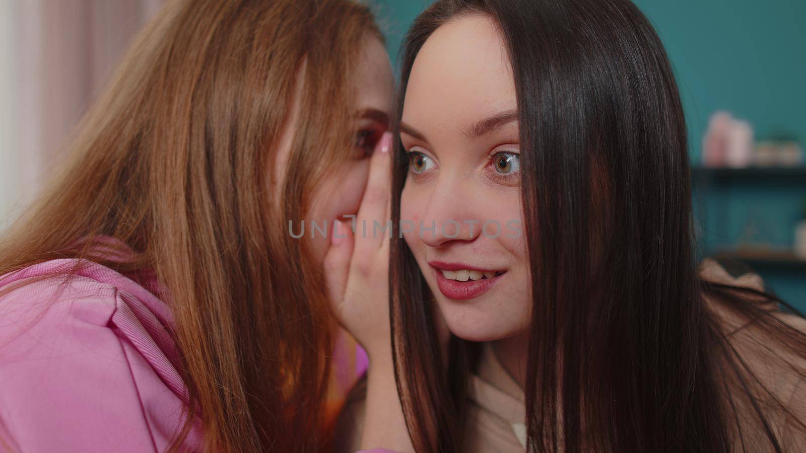 Woman whisper news rumors in ear of excited female girlfriend at home in living room. Young adult smiling girls friends siblings family share secrets gossip having fun laughing together. Close up shot