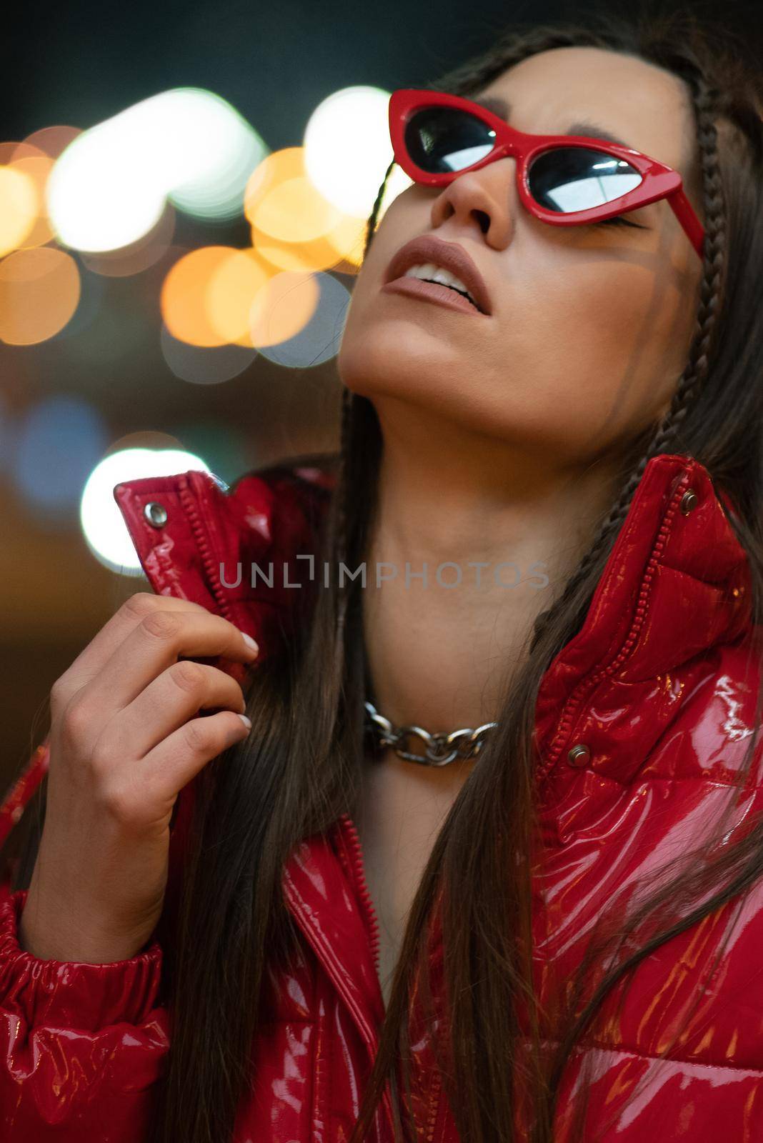 Neon lights night life fashion girl in sun glasses with braided hair and red glossy coat standing outdoors in night city background. Night life concept. Night club life concept.