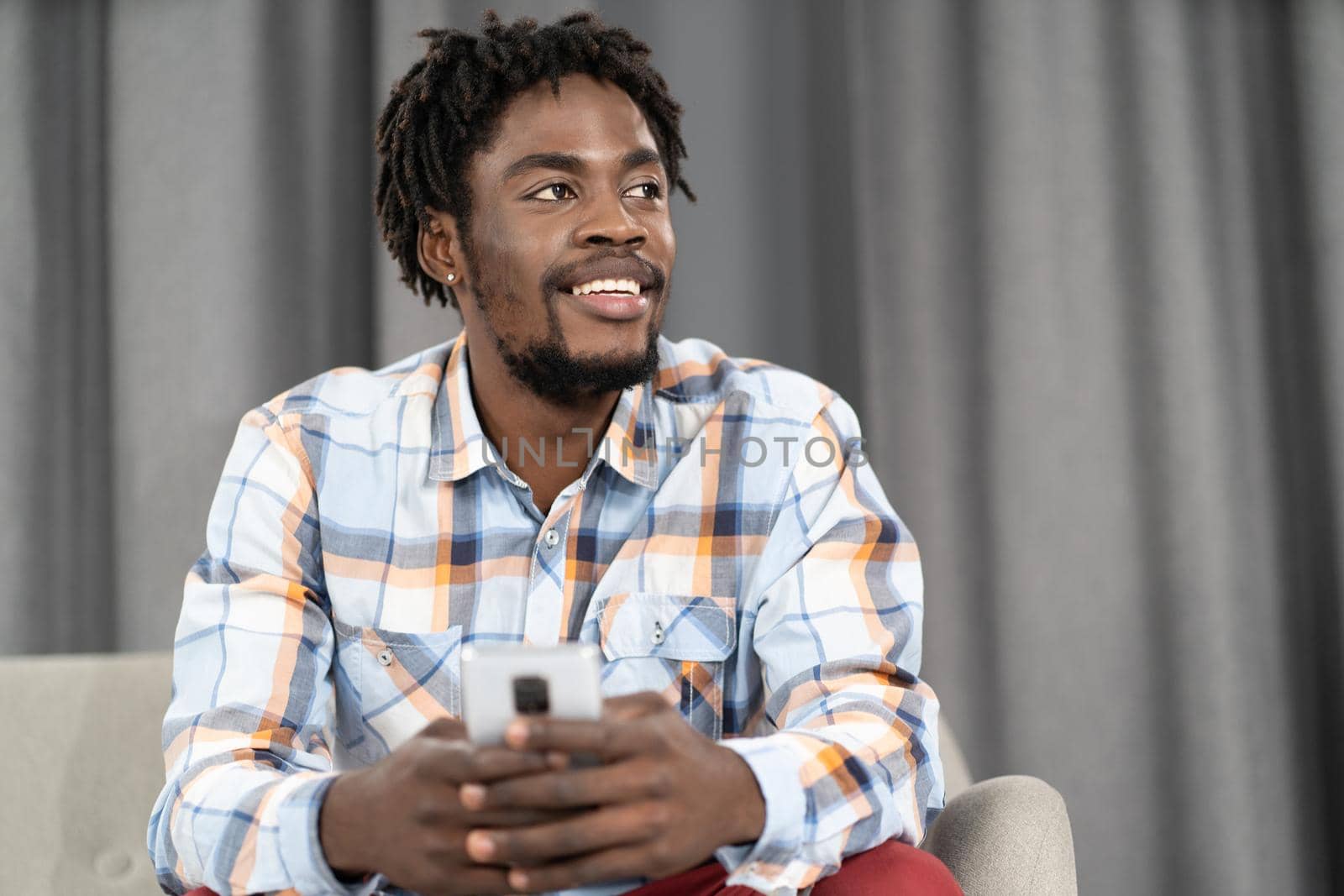 African American young man holding phone or smartphone in hand looking away. Portrait of happy man having present conversation sitting on the sofa. Social media concept.