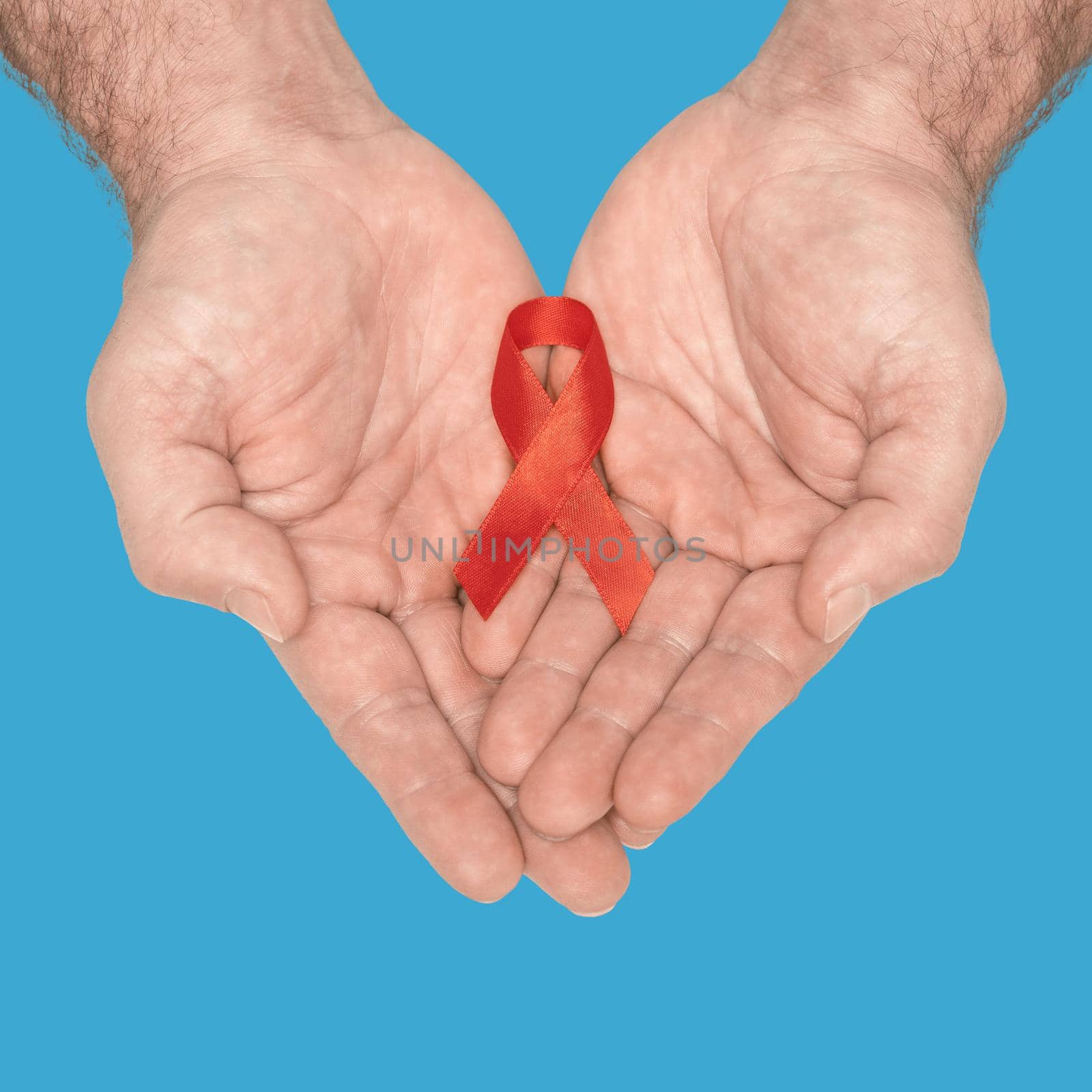 Red awareness ribbon bow on mans helping hands isolated on blue background. HIV, AIDS world day. Social life issues concept. Aids charity fund concept. Healthcare and medicine concept by LipikStockMedia