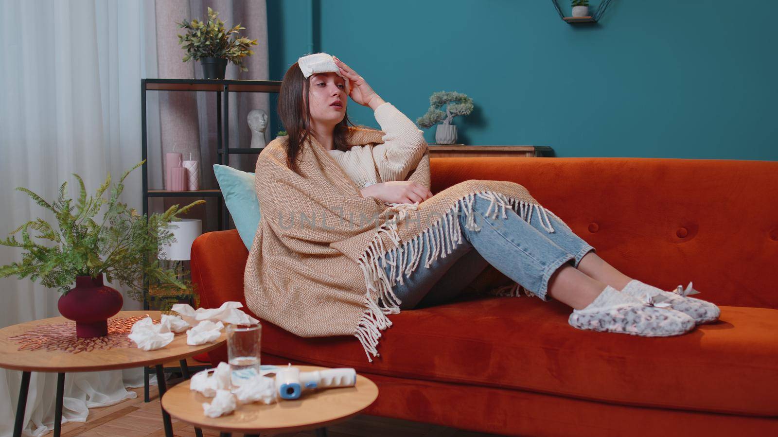 Young one woman suffering from cold or allergy in plaid lying on sofa in living room at home. Sick adult girl with towel on head blows sneezes wipes snot into napkin. Coronavirus quarantine, pandemic