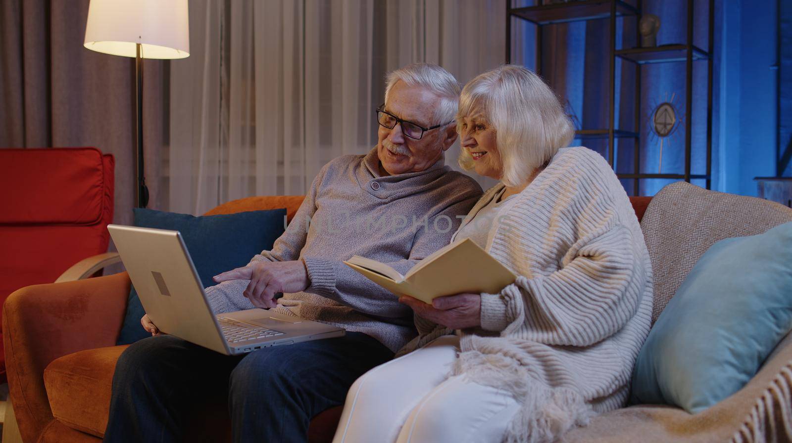 Senior grandparents couple reading book, using laptop pc on couch in night living room at home by efuror