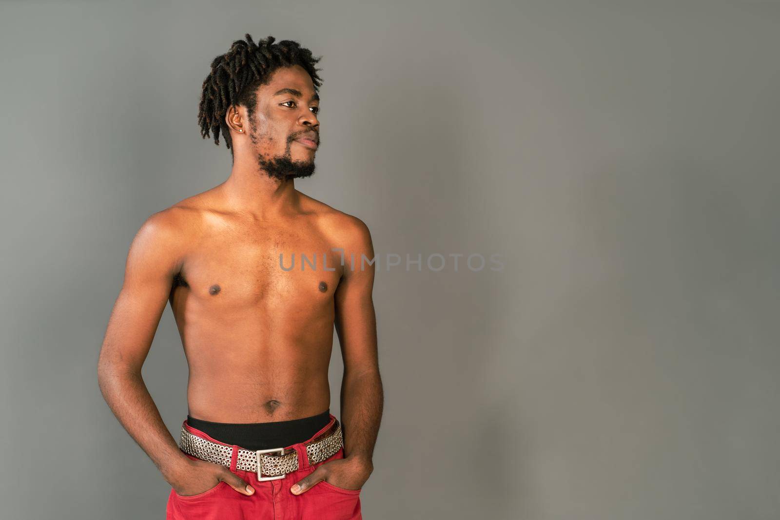 Shirtless african male model standing half turn with hands in pockets. Half naked african american man posing on camera isolated on grey background. Topless african american man. Copy space.