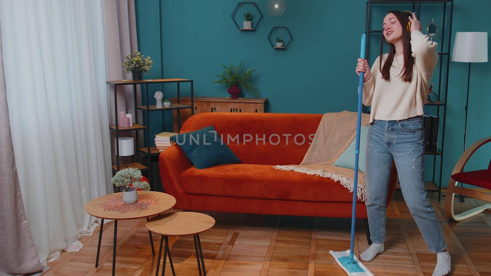 Joyful young woman is mopping floor in light apartment living room, having fun listening to music through headphones, dancing. Girl singing into mop like into a microphone at home. People relaxation
