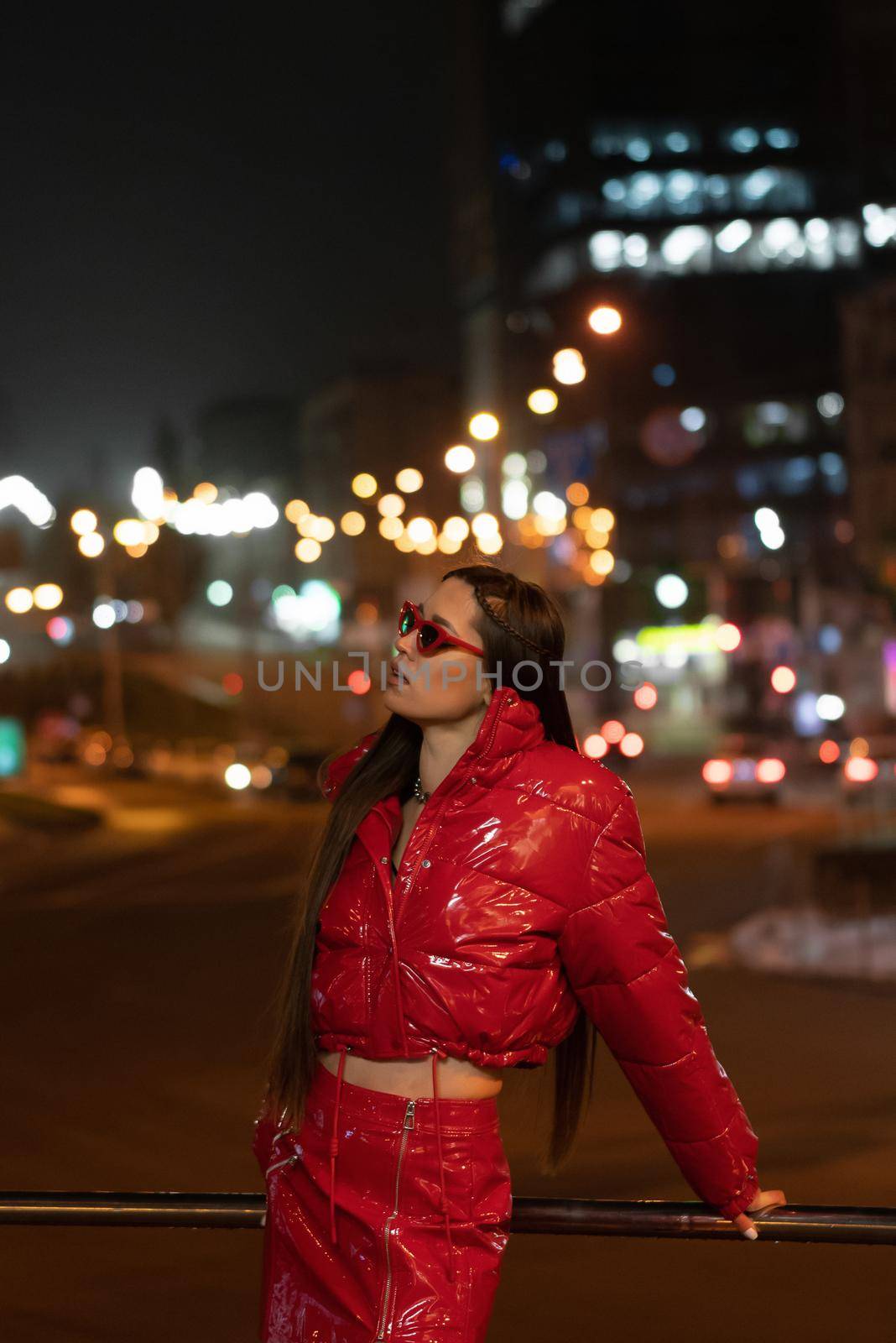 Fashion model girl with afri braid and red shiny skirt and winter coat standing outdoors in night city background. Night life concept. Night club life concept.