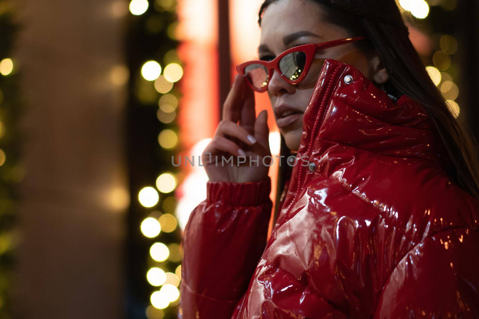 Model fashion girl in sun glasses with braided hair and red glossy coat standing outdoors in night city background. Night life concept. Night club life concept.