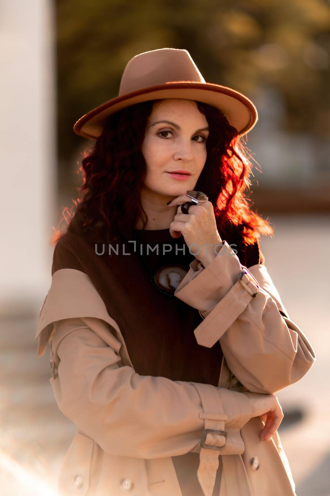 Outdoor fashion portrait of young elegant fashionable brunette woman, model in stylish hat, choker and light raincoat posing at sunset in European city