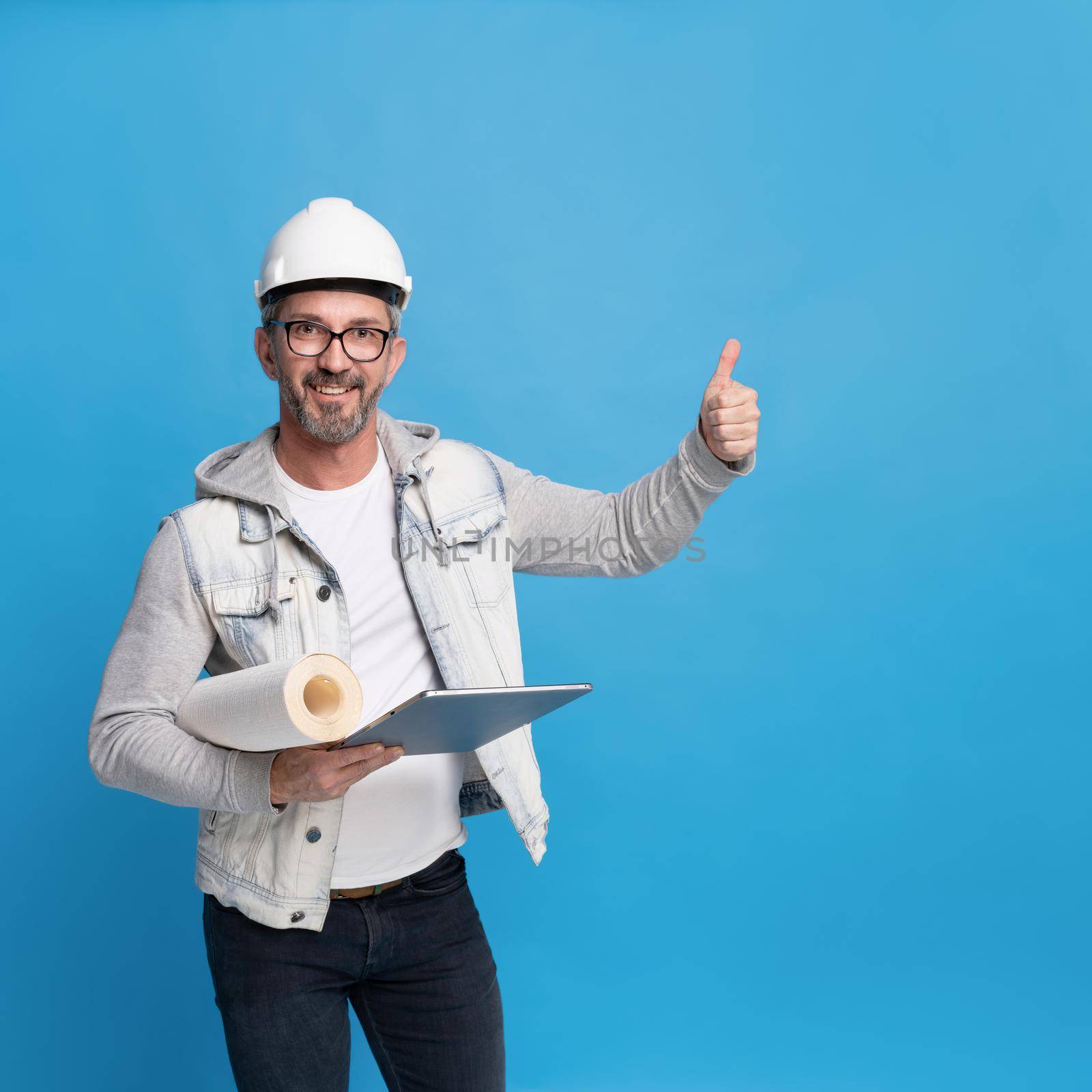Middle aged handsome architecture man holding roll of wall paper and digital tablet pc in trendy casual denim outfit and hard hat or helmet gesturing thumb up or perfect isolated on blue background by LipikStockMedia