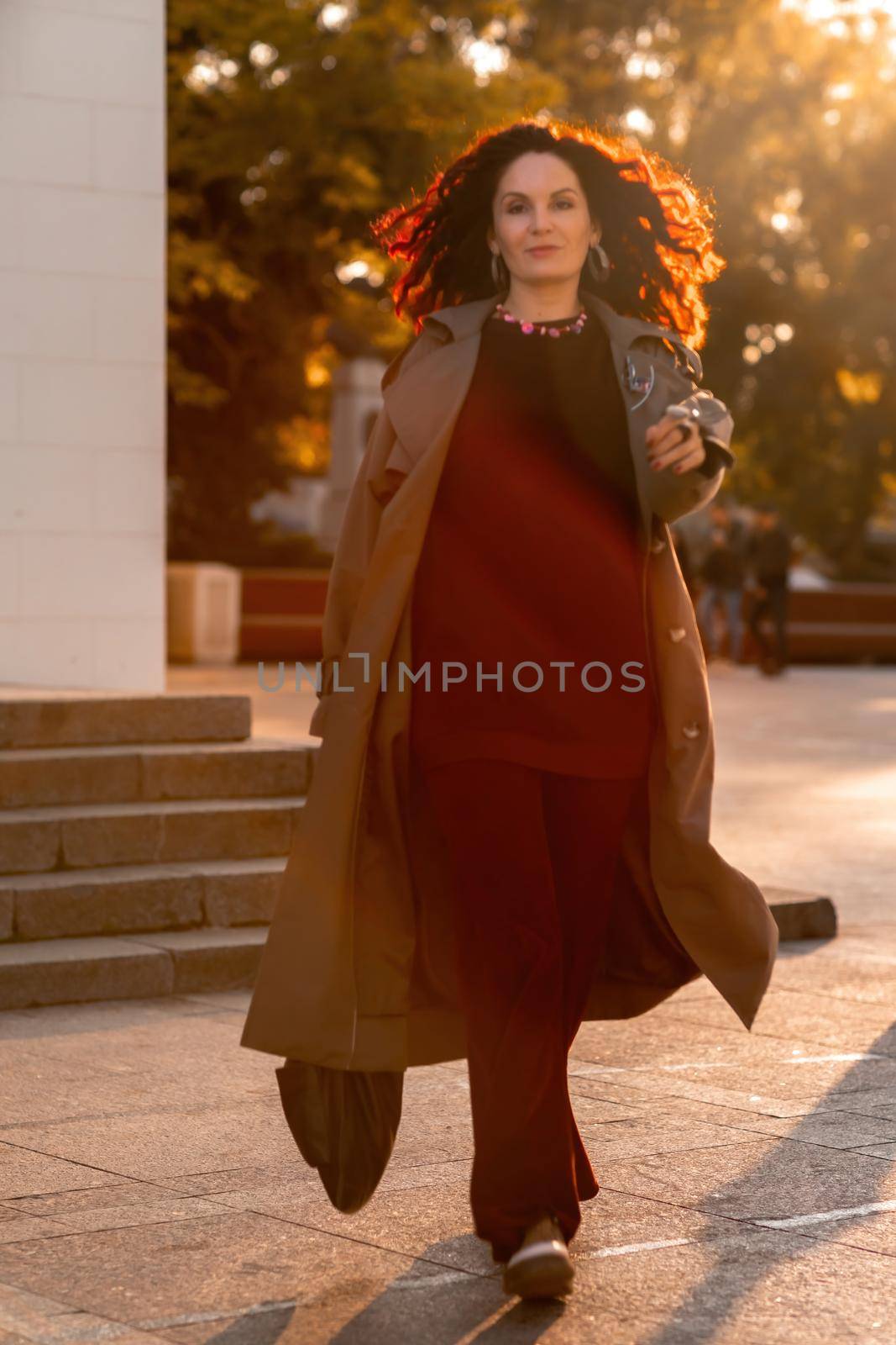 Outdoor fashion portrait of young elegant fashionable brunette woman, model in stylish hat, choker and light raincoat posing at sunset in European city