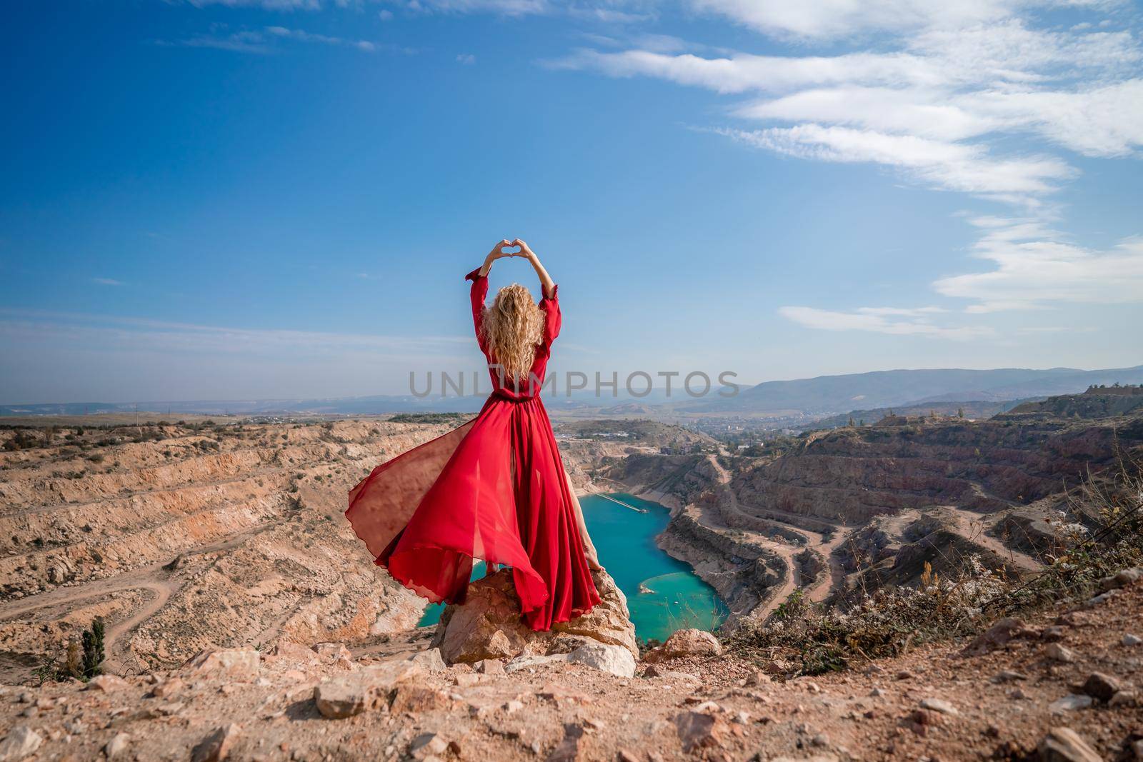 Rear view of a beautiful sensual woman in a red long dress posing on a rock high above the lake in the afternoon. Against the background of the blue sky and the lake in the shape of a heart.