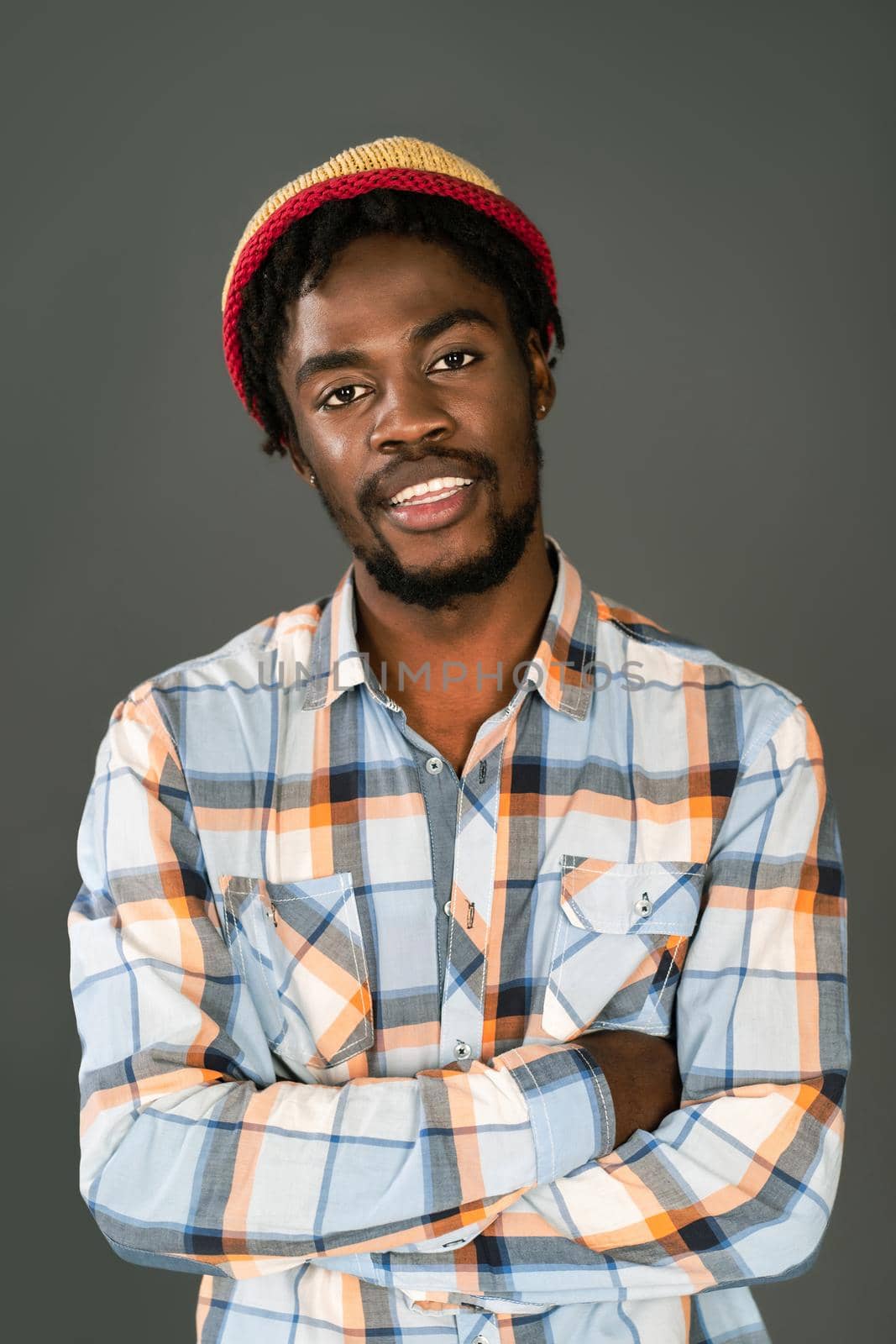 Young smiling black man in afro hat standing with arms folded looking at camera. Cheerful african american young man in plaid shirt looking at camera isolated on gray background by LipikStockMedia