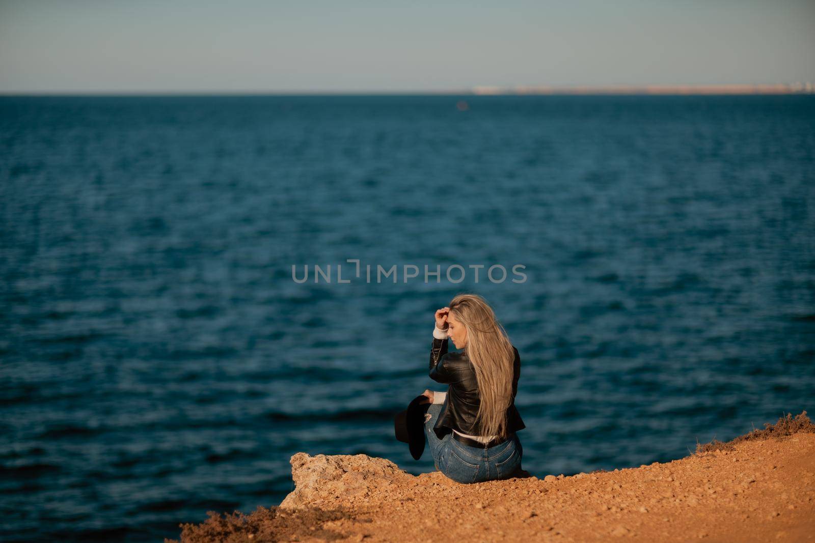 A blonde girl in a stylish black leather jacket is sitting with her back to the seashore