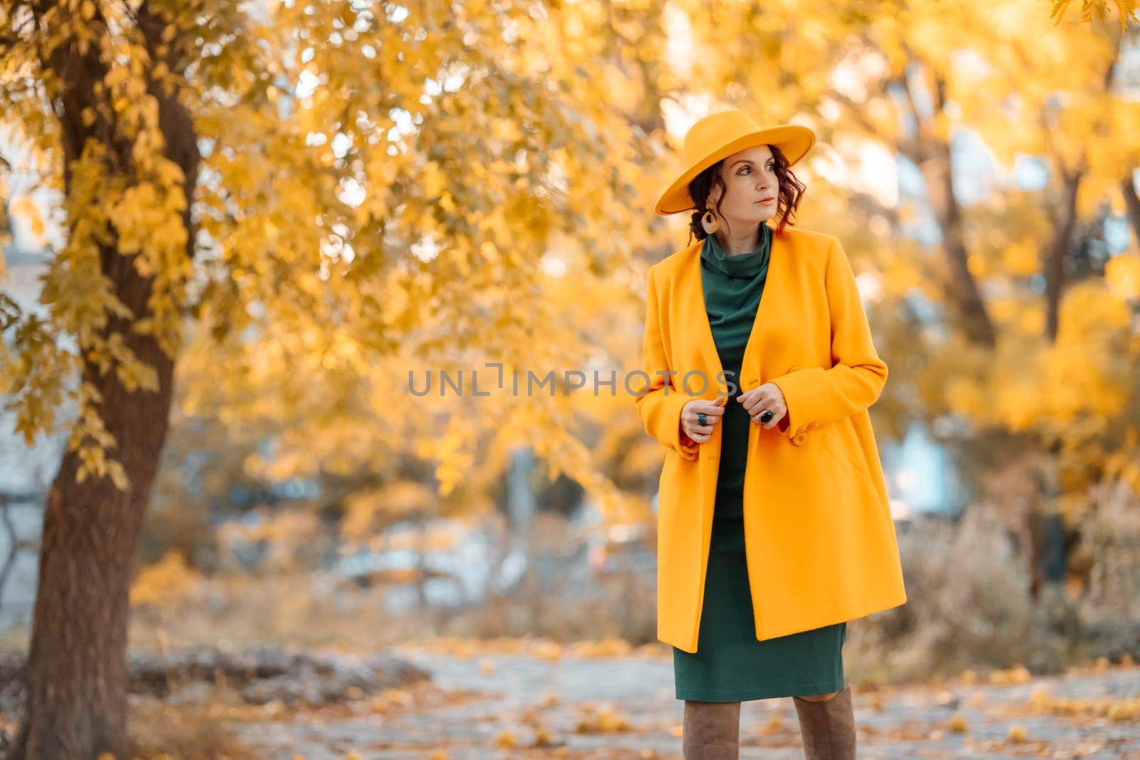 Beautiful woman walks outdoors in autumn. She is wearing a yellow coat, yellow hat and green dress. Young woman enjoying the autumn weather. Autumn content by Matiunina