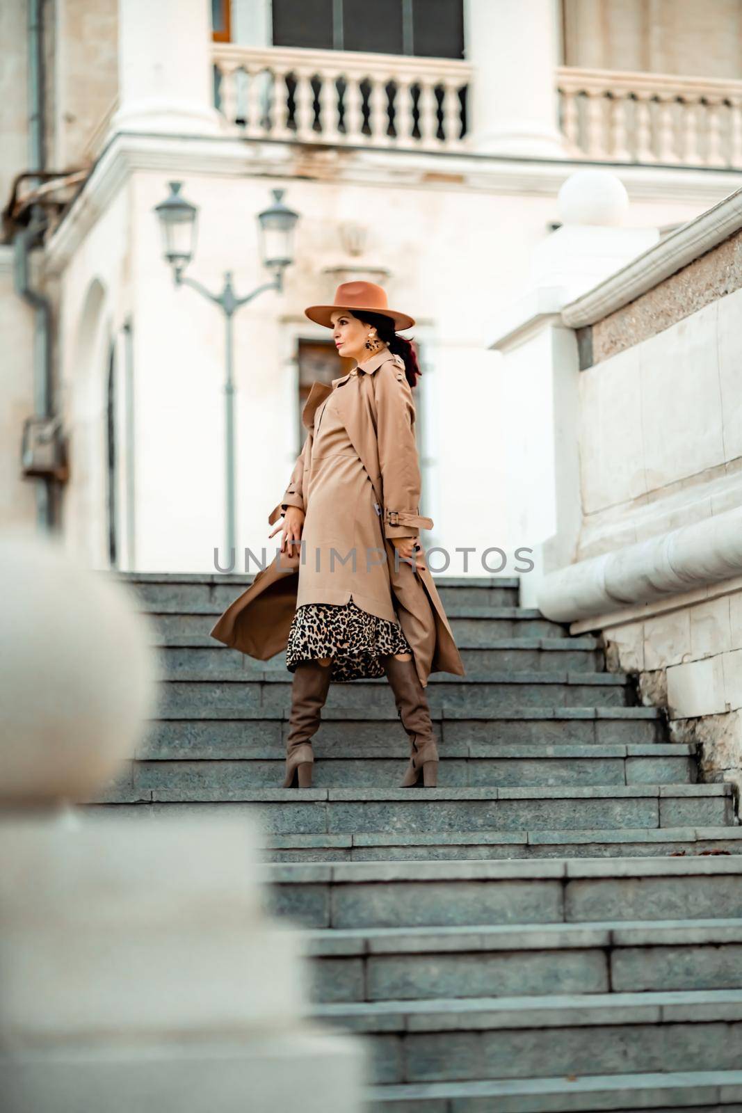Outdoor fashion portrait of young elegant fashionable brunette woman, model in stylish hat, choker and light raincoat posing at sunset in European city