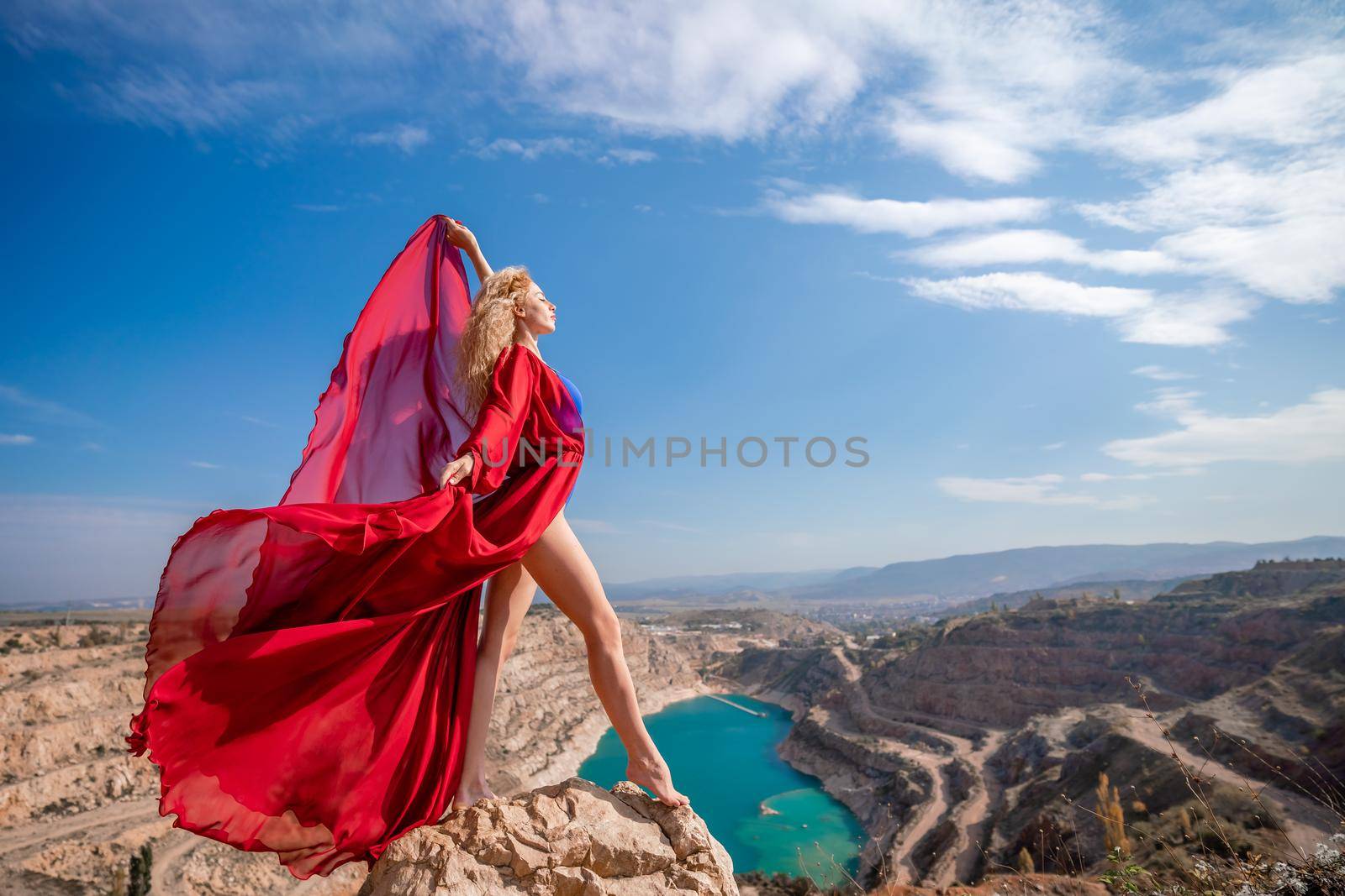 Side view of a beautiful sensual woman in a red long dress posing on a rock high above the lake in the afternoon. Against the background of the blue sky and the lake in the form of a heart.