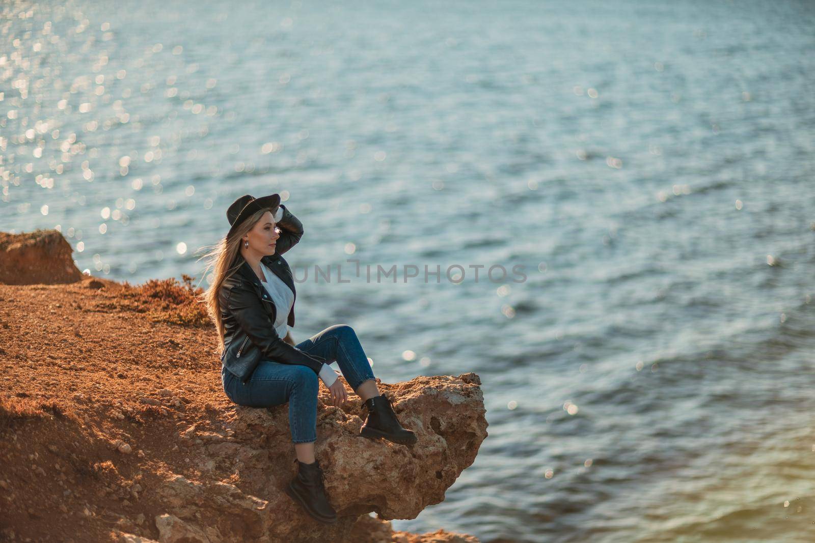 A blonde girl in a stylish black leather jacket is sitting on the seashore