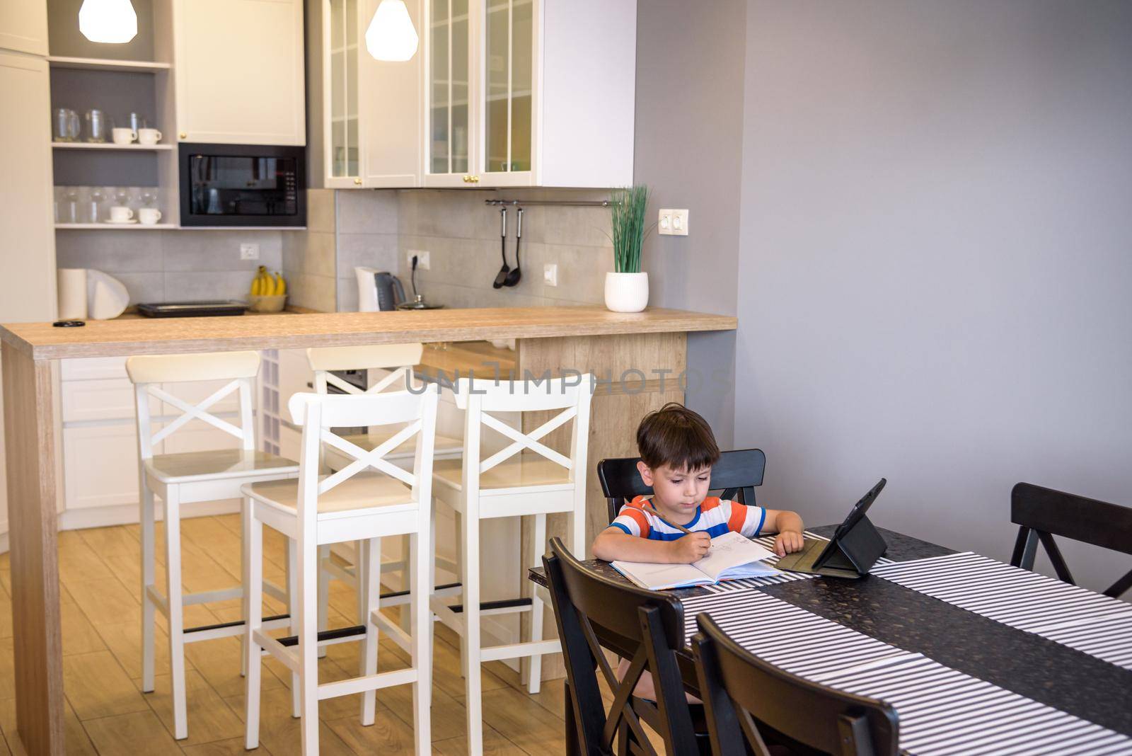Smart preteen schoolboy doing his homework with digital tablet at home. Child using gadgets on his kitchen to study. Modern education and learning for kids.