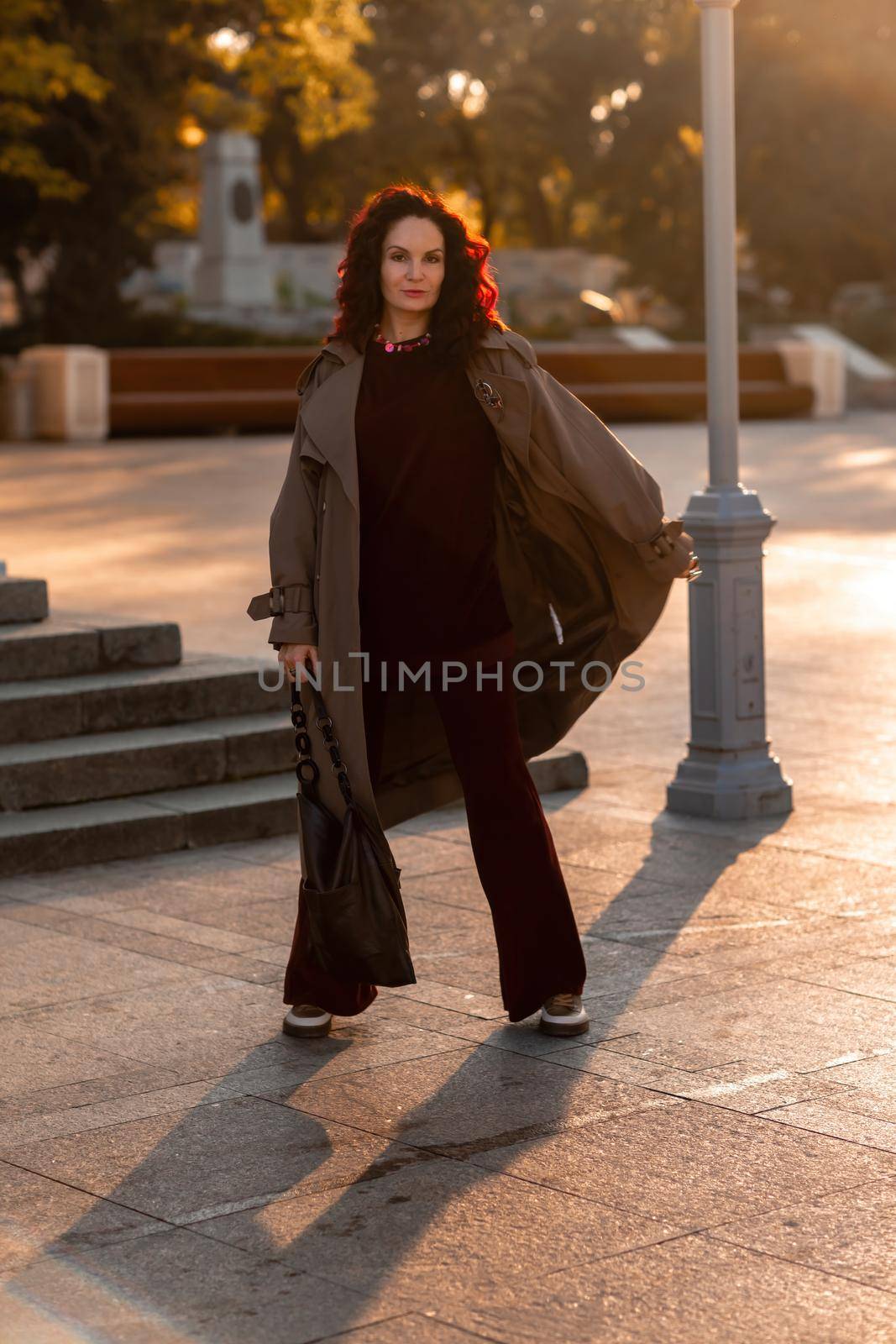 Outdoor fashion portrait of young elegant fashionable brunette woman, model in stylish hat, choker and light raincoat posing at sunset in European city