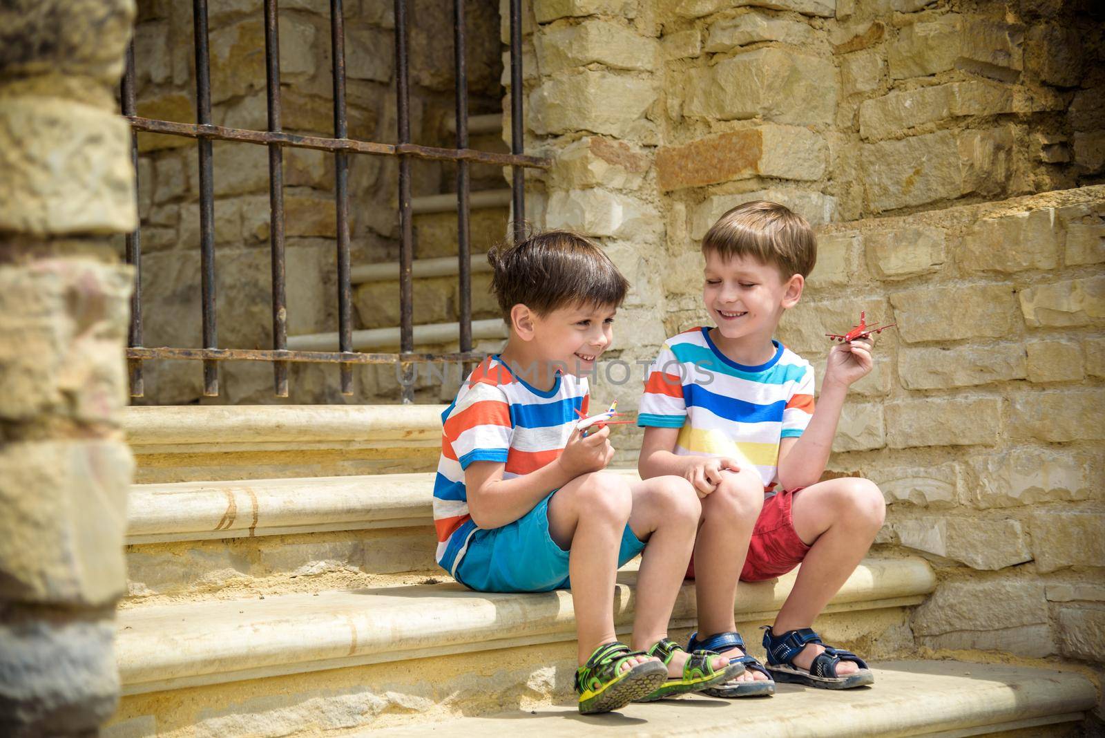 The children playing on the ruins of ancient building with metal gate an archaeological site of an ancient city. Two boys sitting and play with toy aircraft plane. Travel concept.