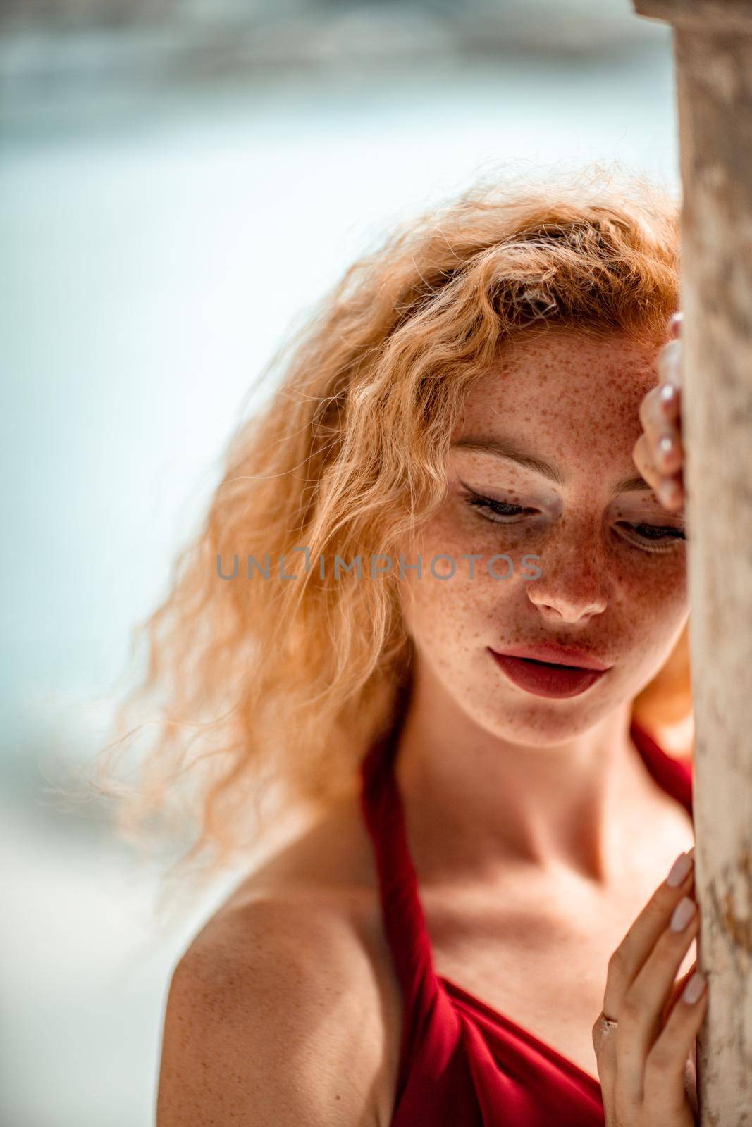 Outdoor portrait of a young beautiful natural redhead girl with freckles, long curly hair, in a red dress, posing against the background of the sea