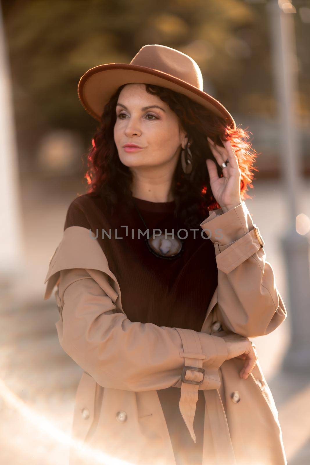 Outdoor fashion portrait of young elegant fashionable brunette woman, model in stylish hat, choker and light raincoat posing at sunset in European city