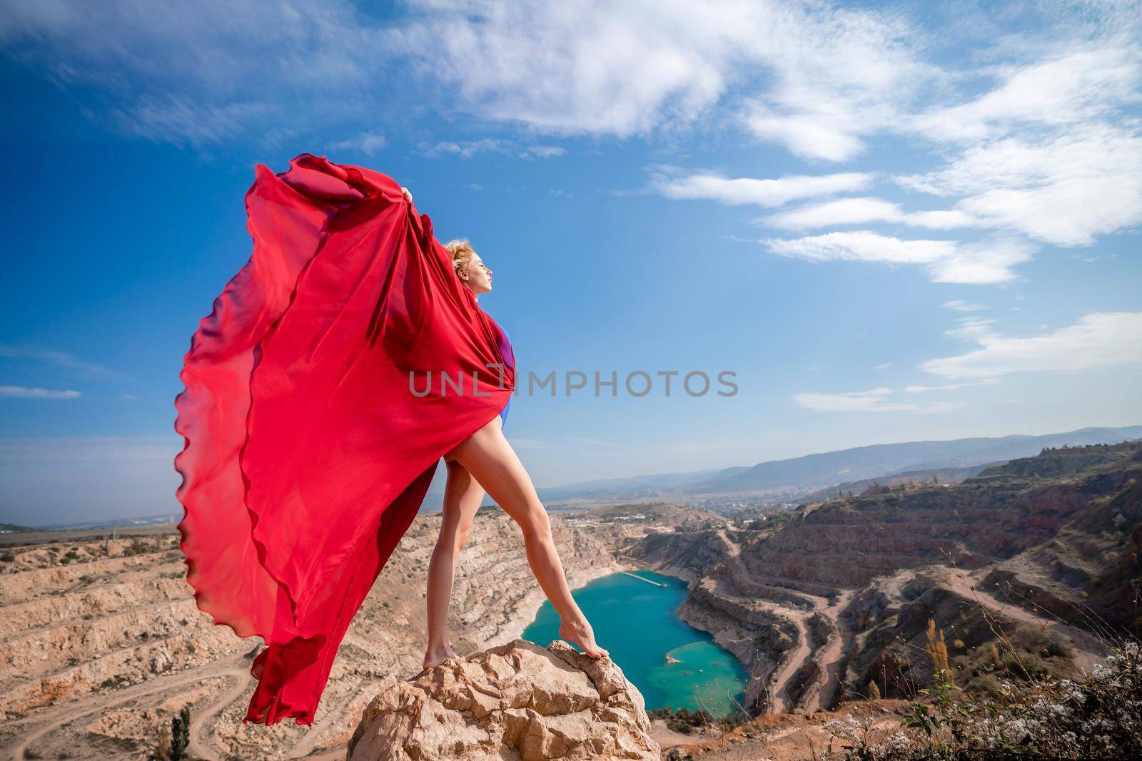 Side view of a beautiful sensual woman in a red long dress posing on a rock high above the lake in the afternoon. Against the background of the blue sky and the lake in the form of a heart.