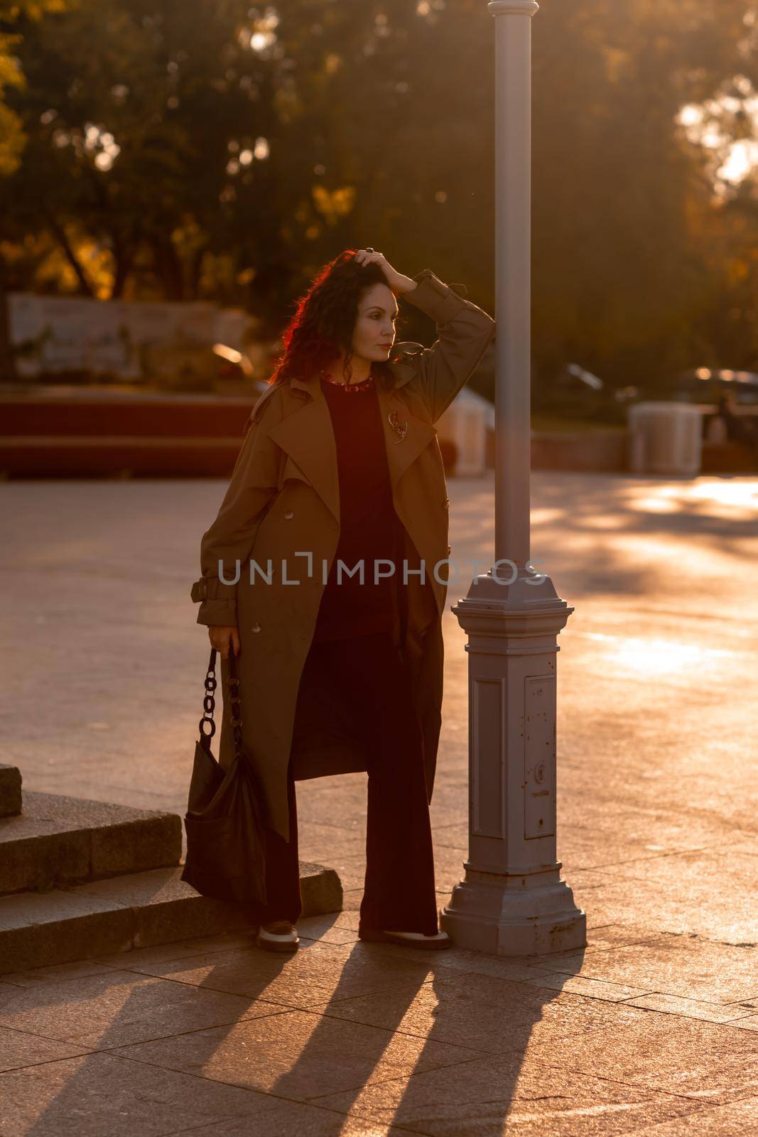 Outdoor fashion portrait of young elegant fashionable brunette woman, model in stylish hat, choker and light raincoat posing at sunset in European city