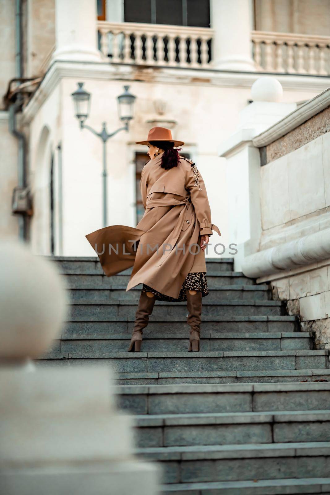 Outdoor fashion portrait of young elegant fashionable brunette woman, model in stylish hat, choker and light raincoat posing at sunset in European city