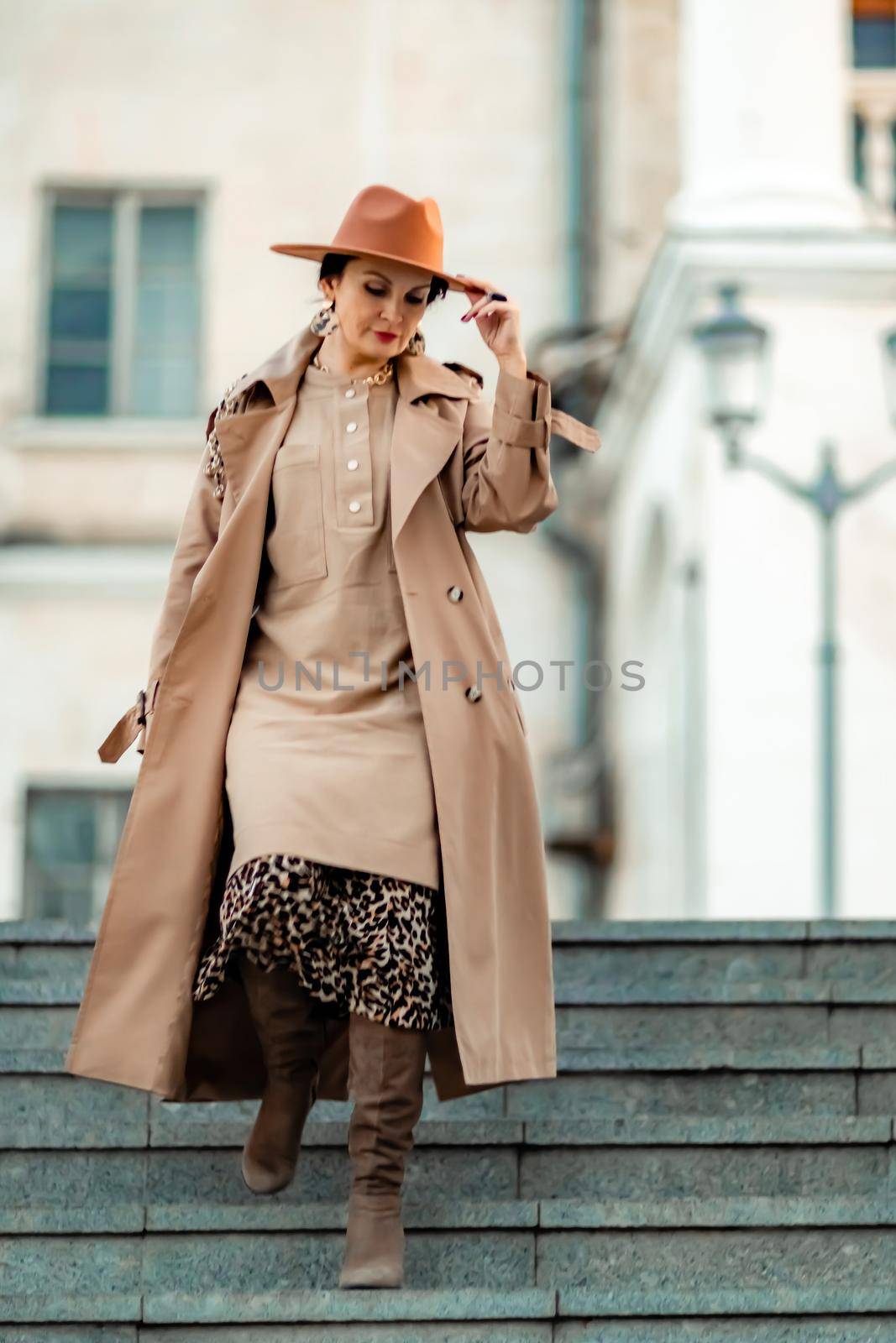 Outdoor fashion portrait of young elegant fashionable brunette woman, model in stylish hat, choker and light raincoat posing at sunset in European city