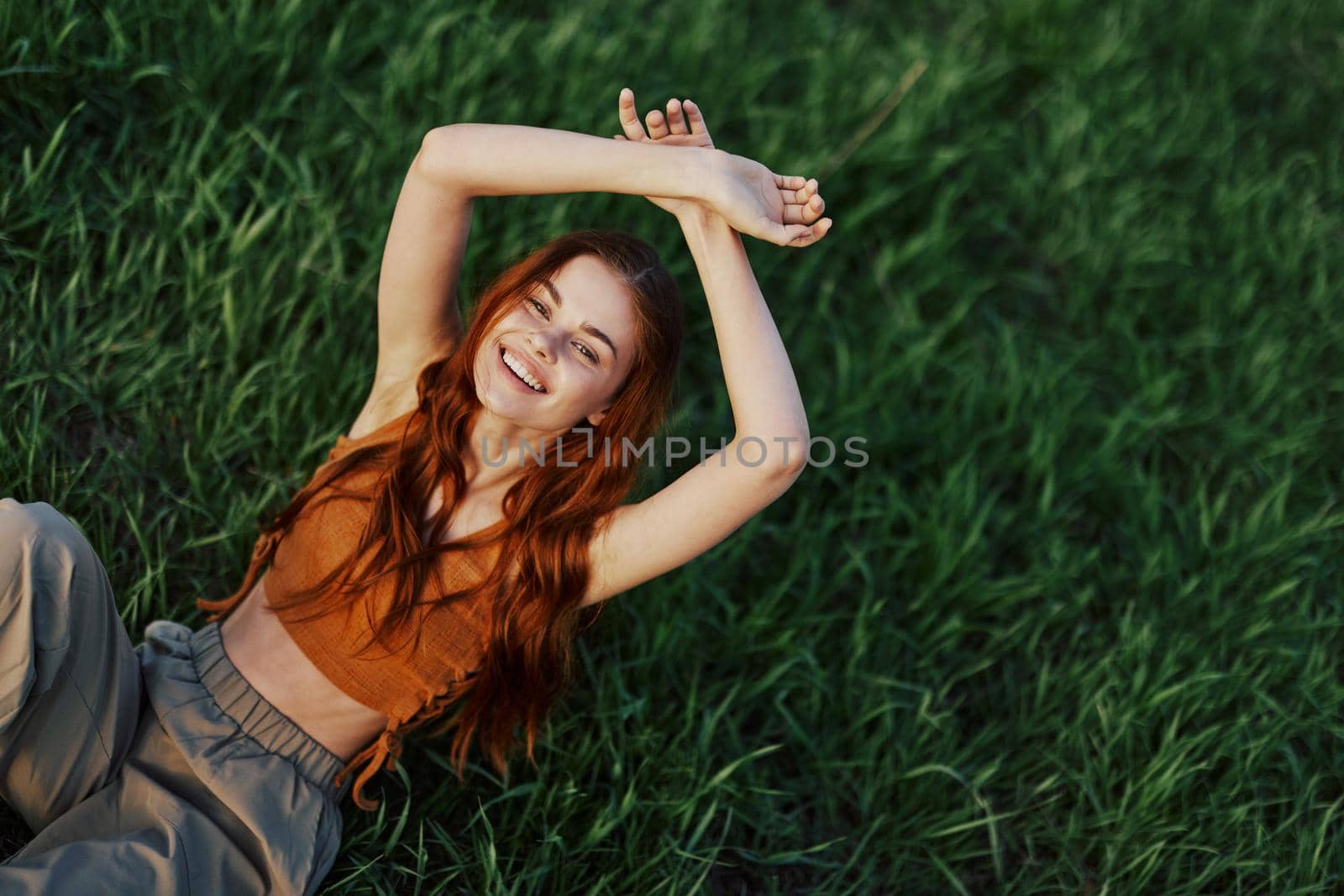 A woman lies in nature on the green grass in the park resting with her hands up and smiling, her red hair flying in the wind in the open air in the sunlight. Freelancer's concept on vacation. High quality photo