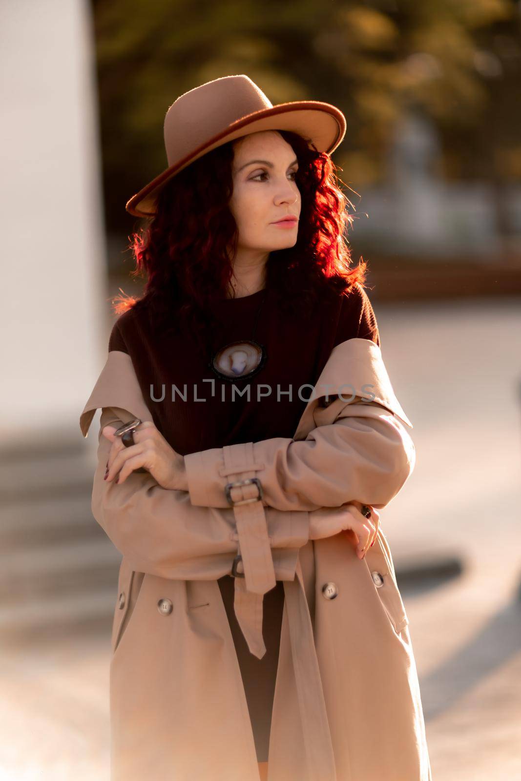 Outdoor fashion portrait of young elegant fashionable brunette woman, model in stylish hat, choker and light raincoat posing at sunset in European city