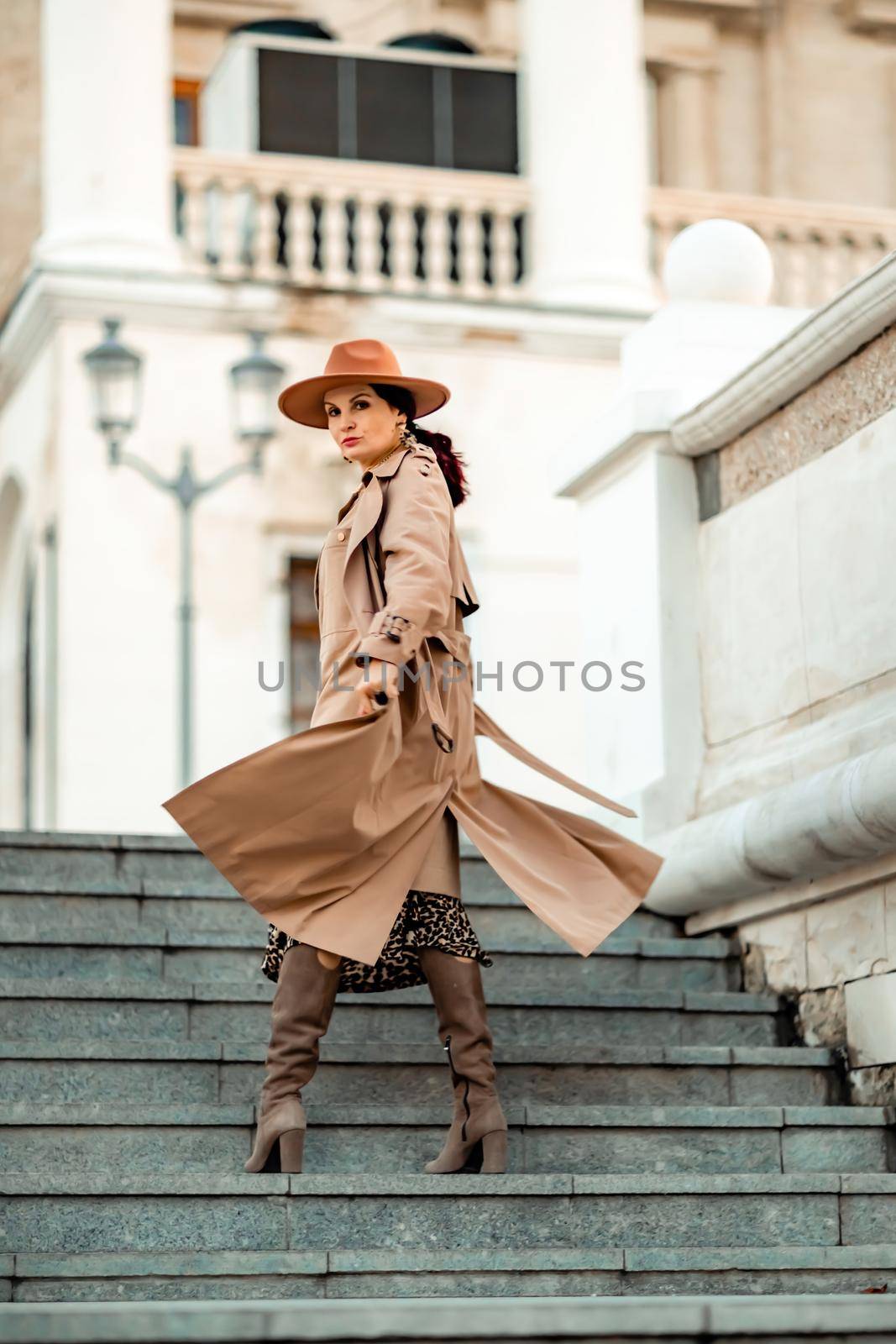 Outdoor fashion portrait of young elegant fashionable brunette woman, model in stylish hat, choker and light raincoat posing at sunset in European city