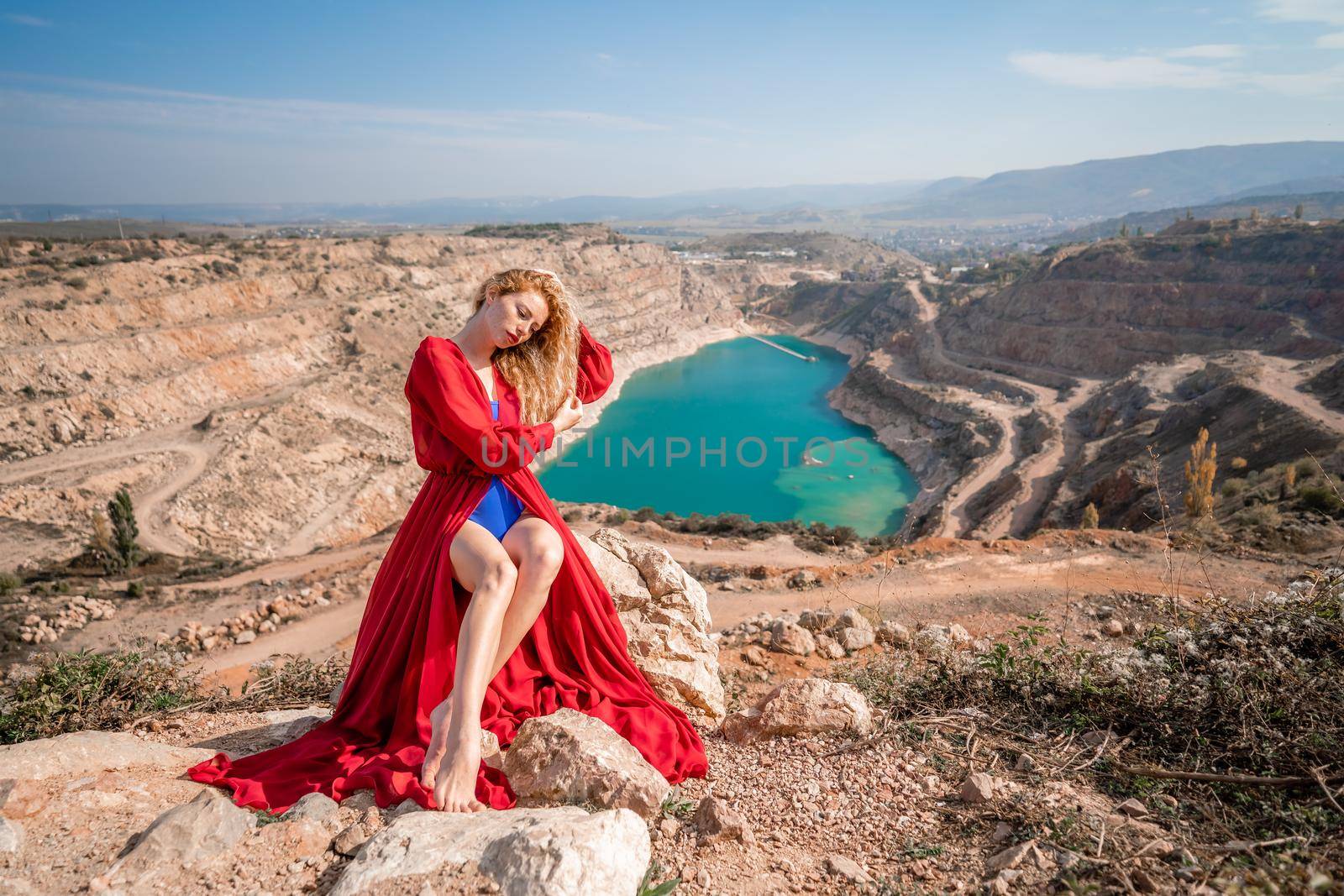 A beautiful girl in a red long dress, Sits on a rock high above the lake in the afternoon. Against the background of the blue sky and the lake in the shape of a heart. by Matiunina